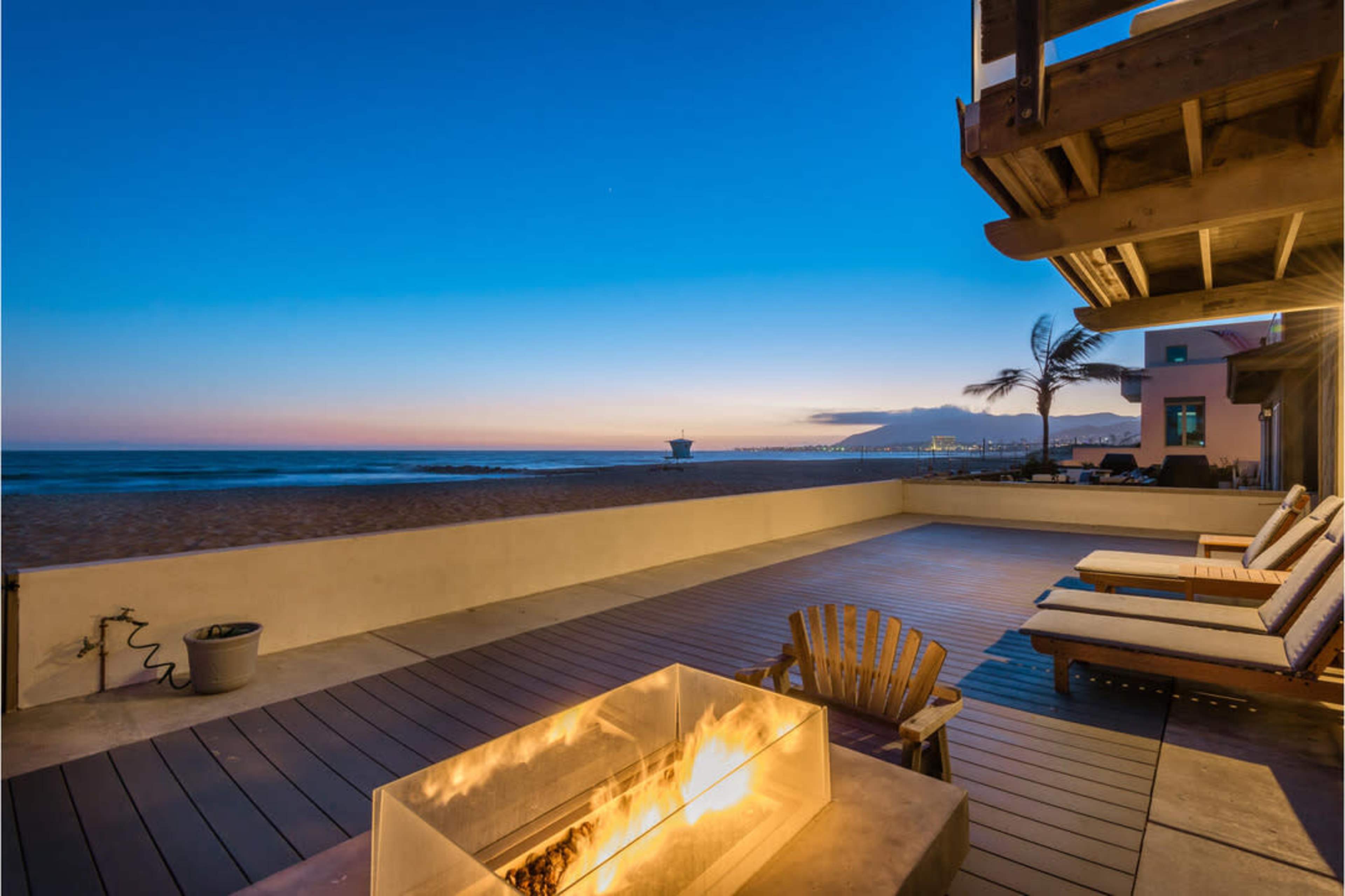 A patio with lounge chairs and a fire pit overlooks the beach at sunset, with the ocean and distant mountains visible in the background.