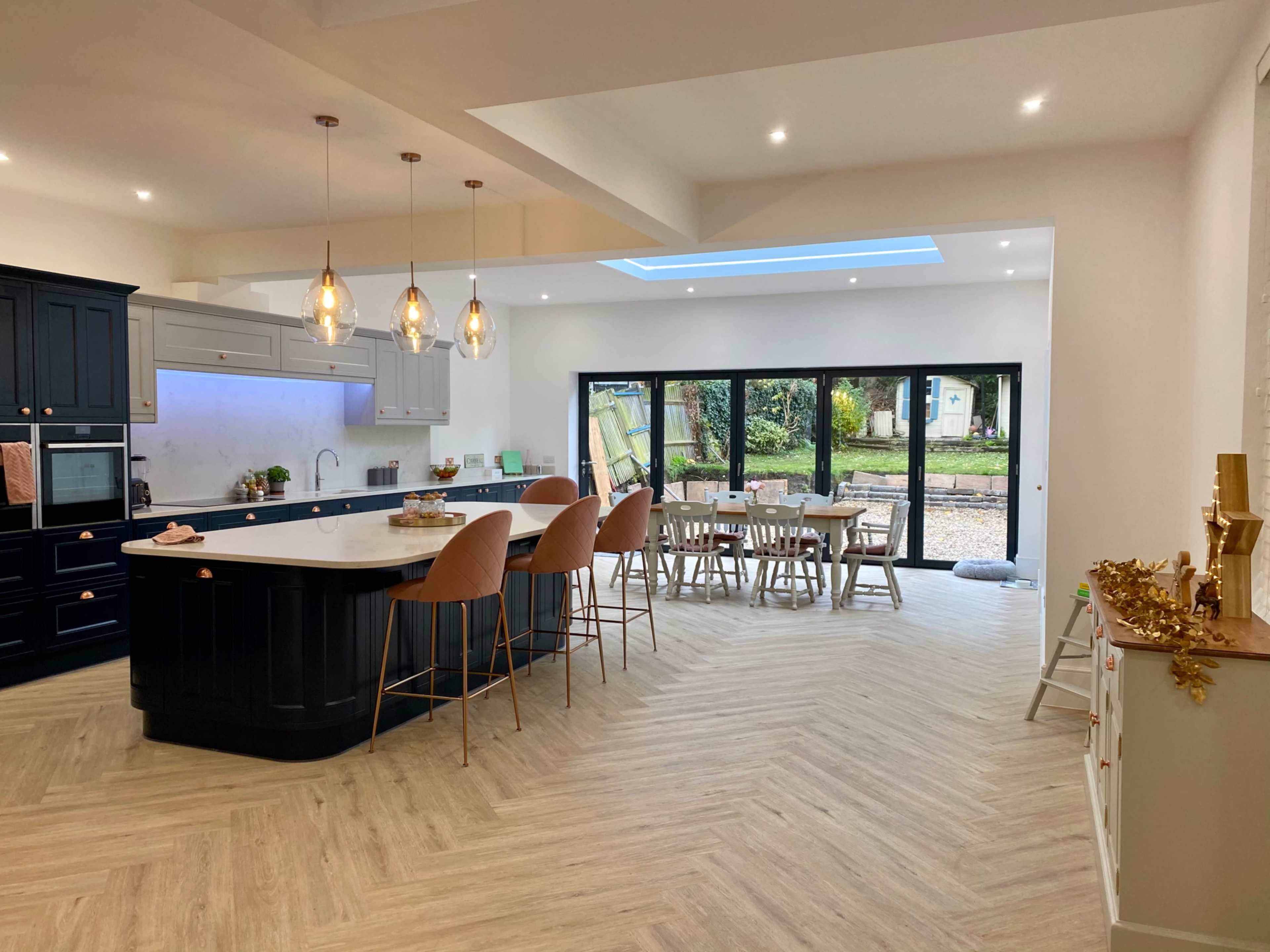 The image shows a modern kitchen and dining area with an island, bar stools, and large glass doors leading to an outdoor space.