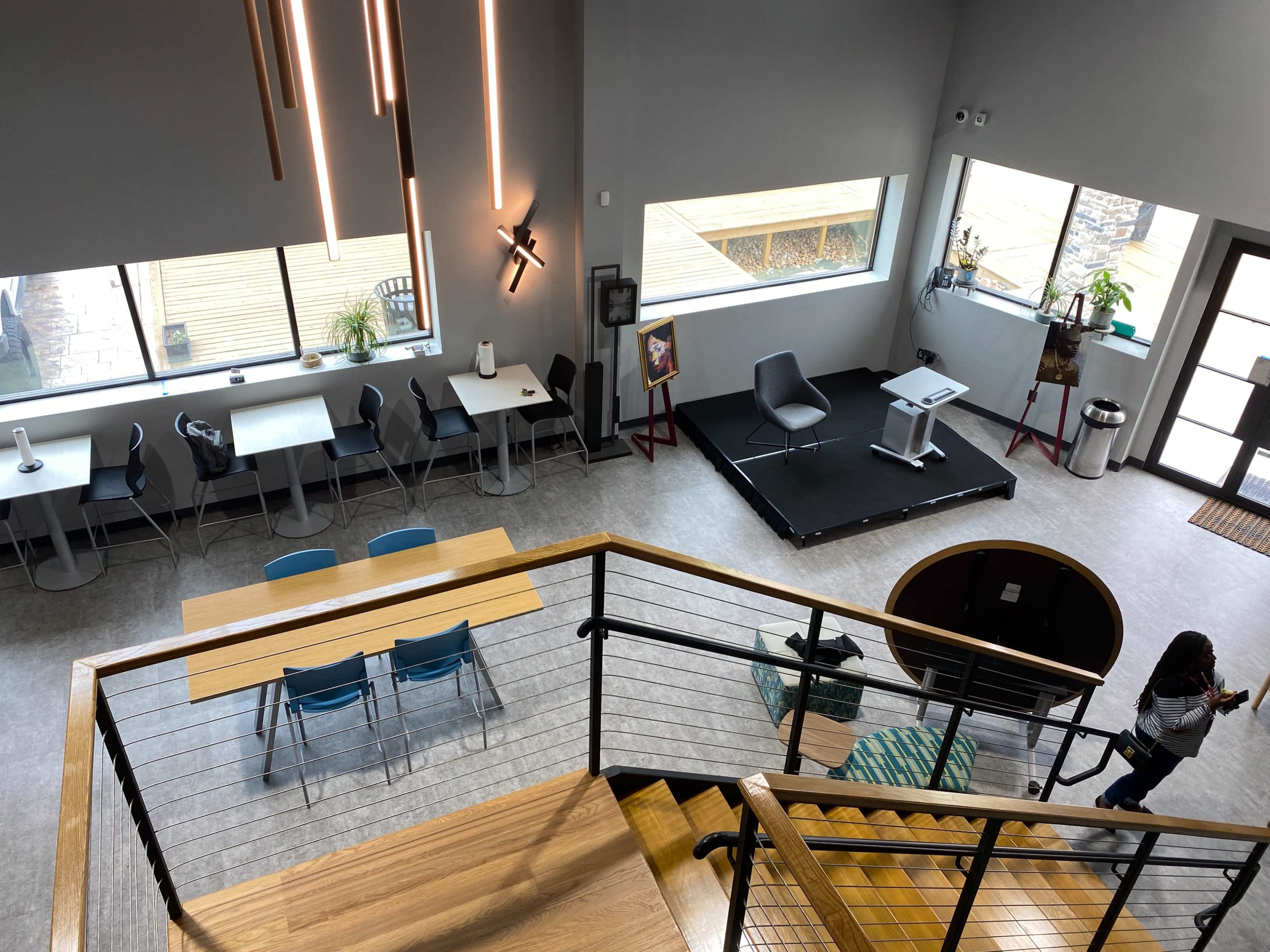The image shows a modern, two-level interior space featuring a dining area with tables and chairs, a stage, and large windows allowing natural light.