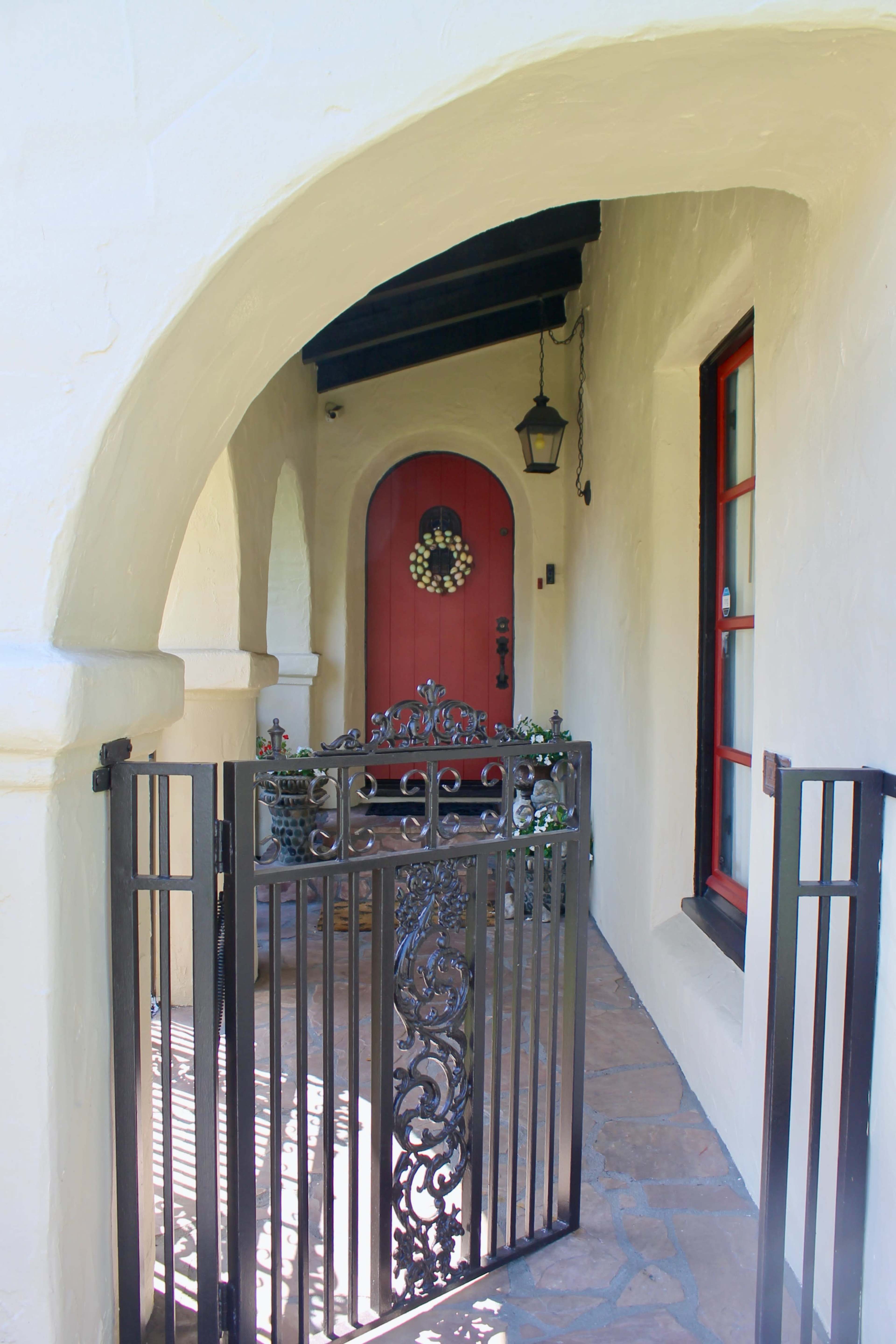 A black wrought iron gate opens to a white stucco entryway featuring a red door adorned with a decorative wreath.
