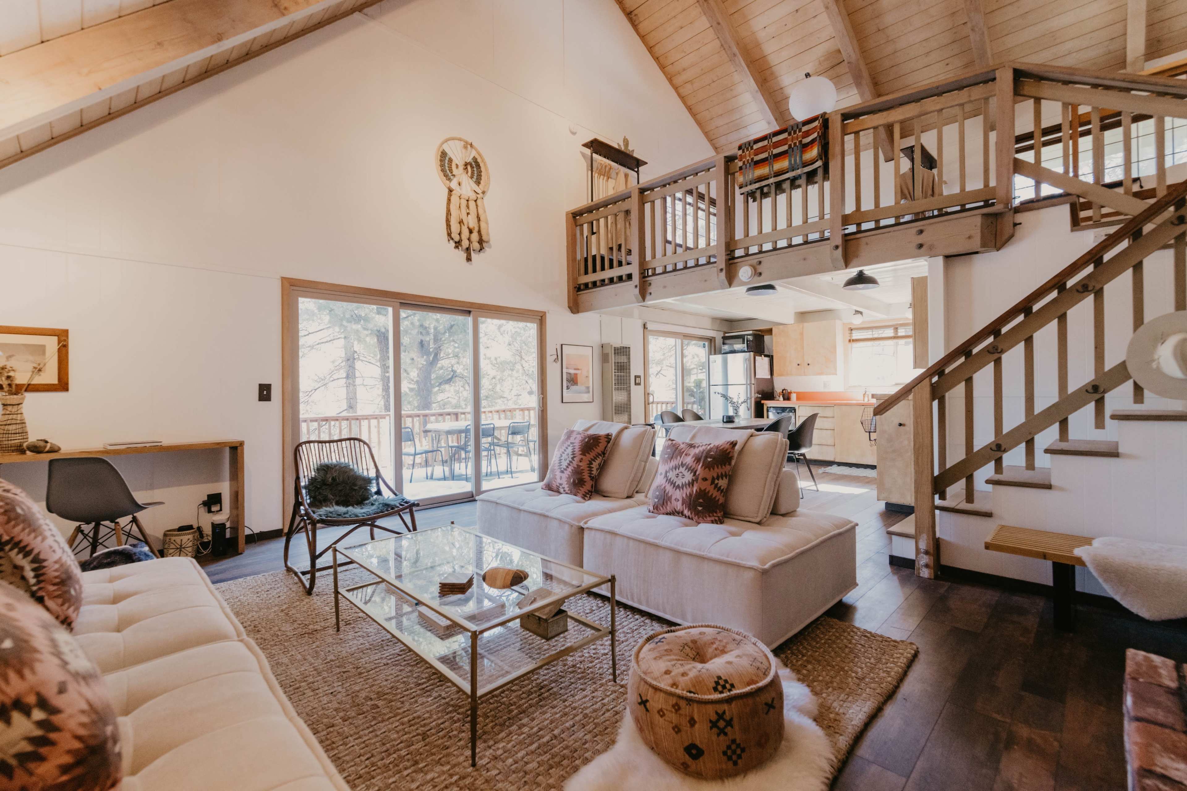 A cozy living room with a sectional sofa, a glass coffee table, and a view of a kitchen and staircase in a cabin-like setting.