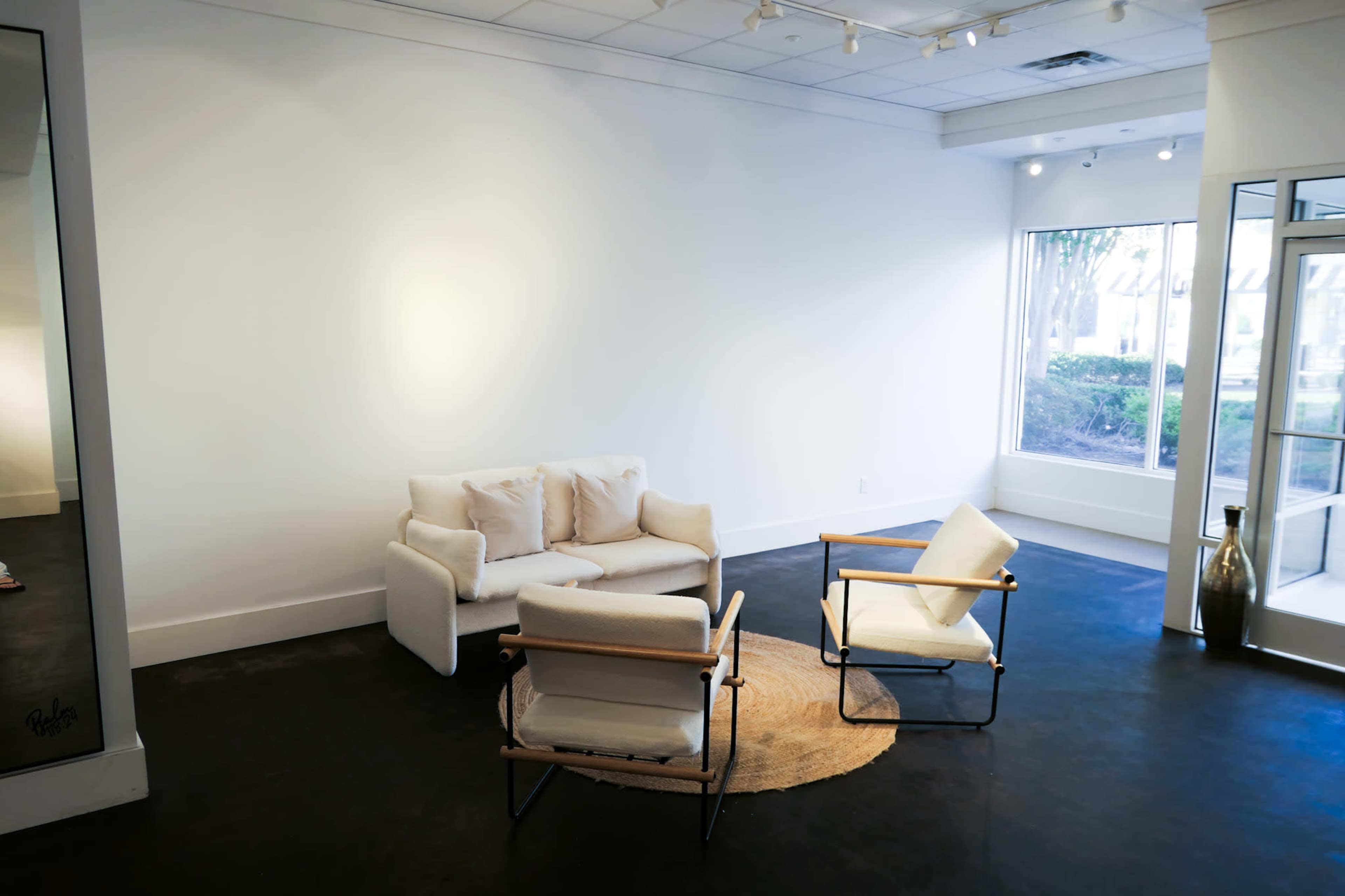 The image shows a minimalist room with white walls, a small white sofa, two chairs, and a round rug, illuminated by natural light from a nearby window.