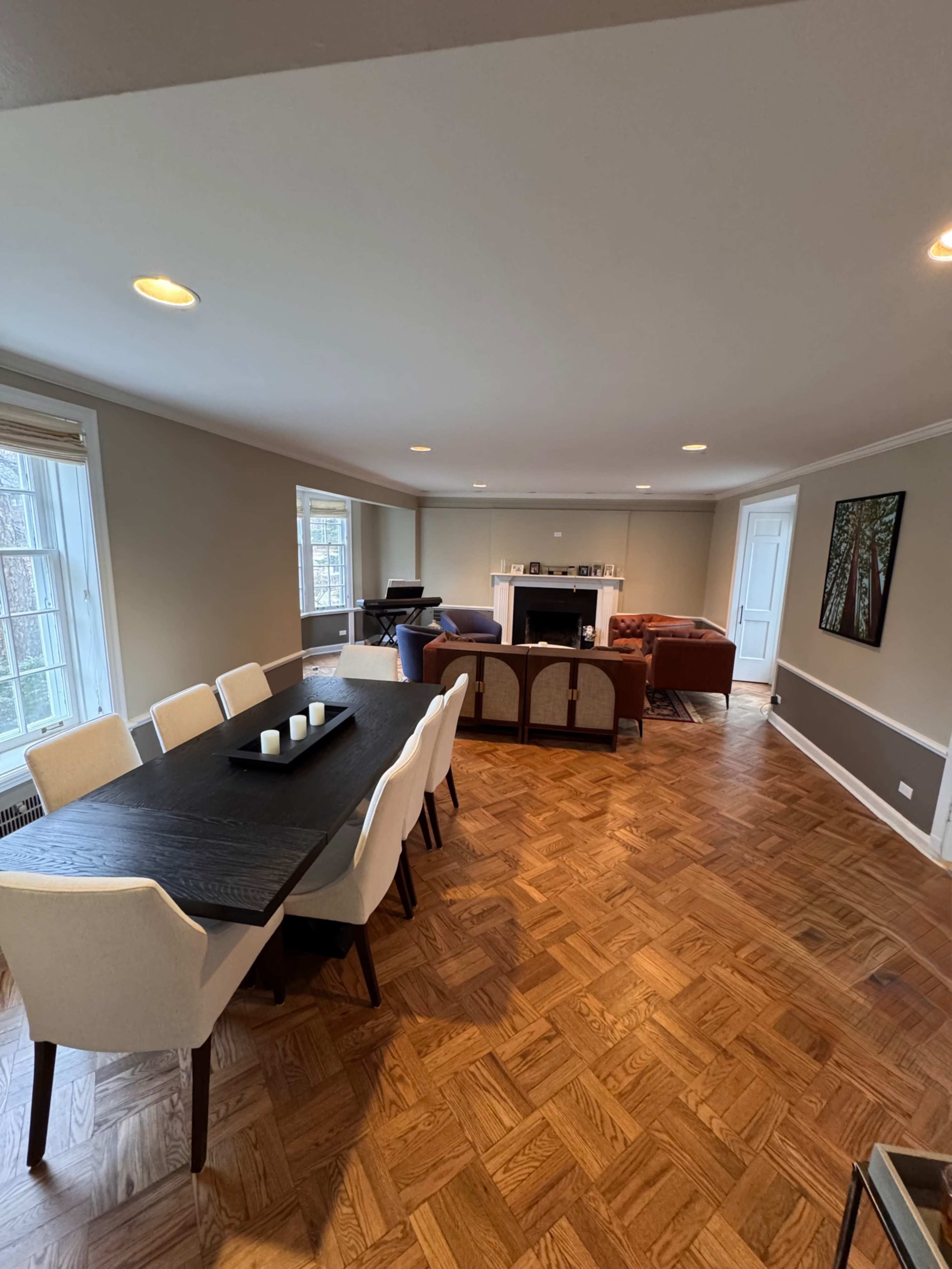 The image shows a spacious living area with a dining table, several seating arrangements, and large windows allowing natural light.