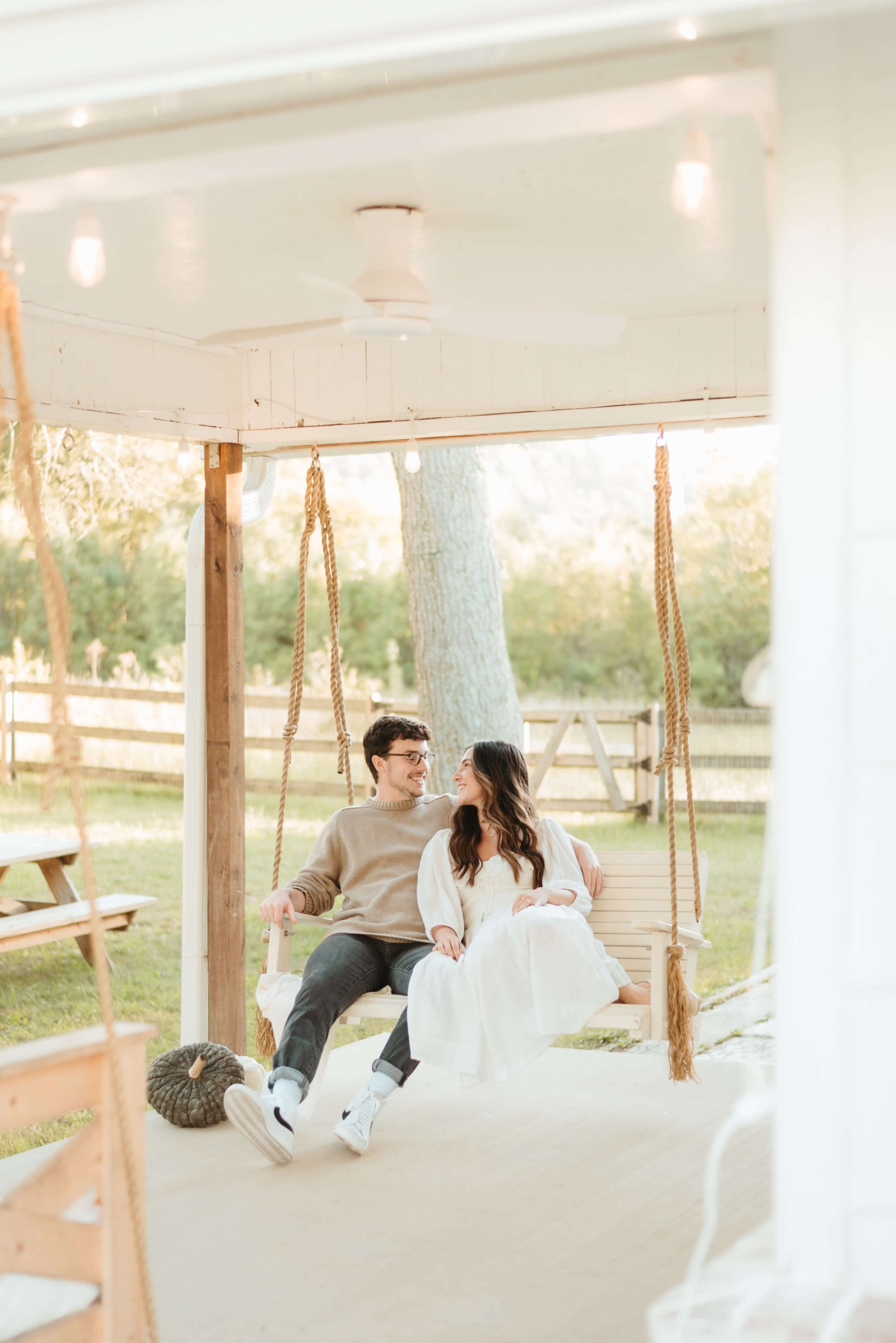 A couple sits together on a swing under a porch, surrounded by greenery and wooden fencing.