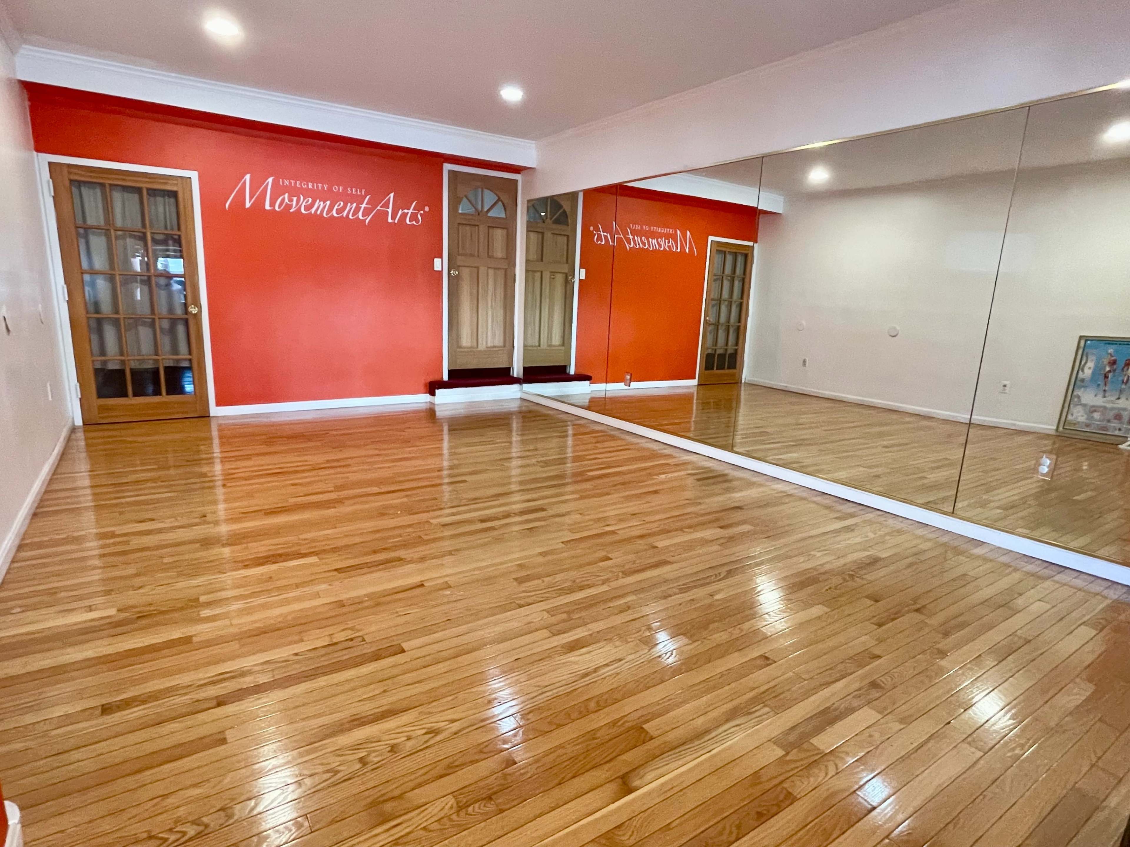 The image shows a spacious room with polished wooden floors, an orange accent wall featuring the text "Movement Arts," and a large mirror reflecting the room's interior.