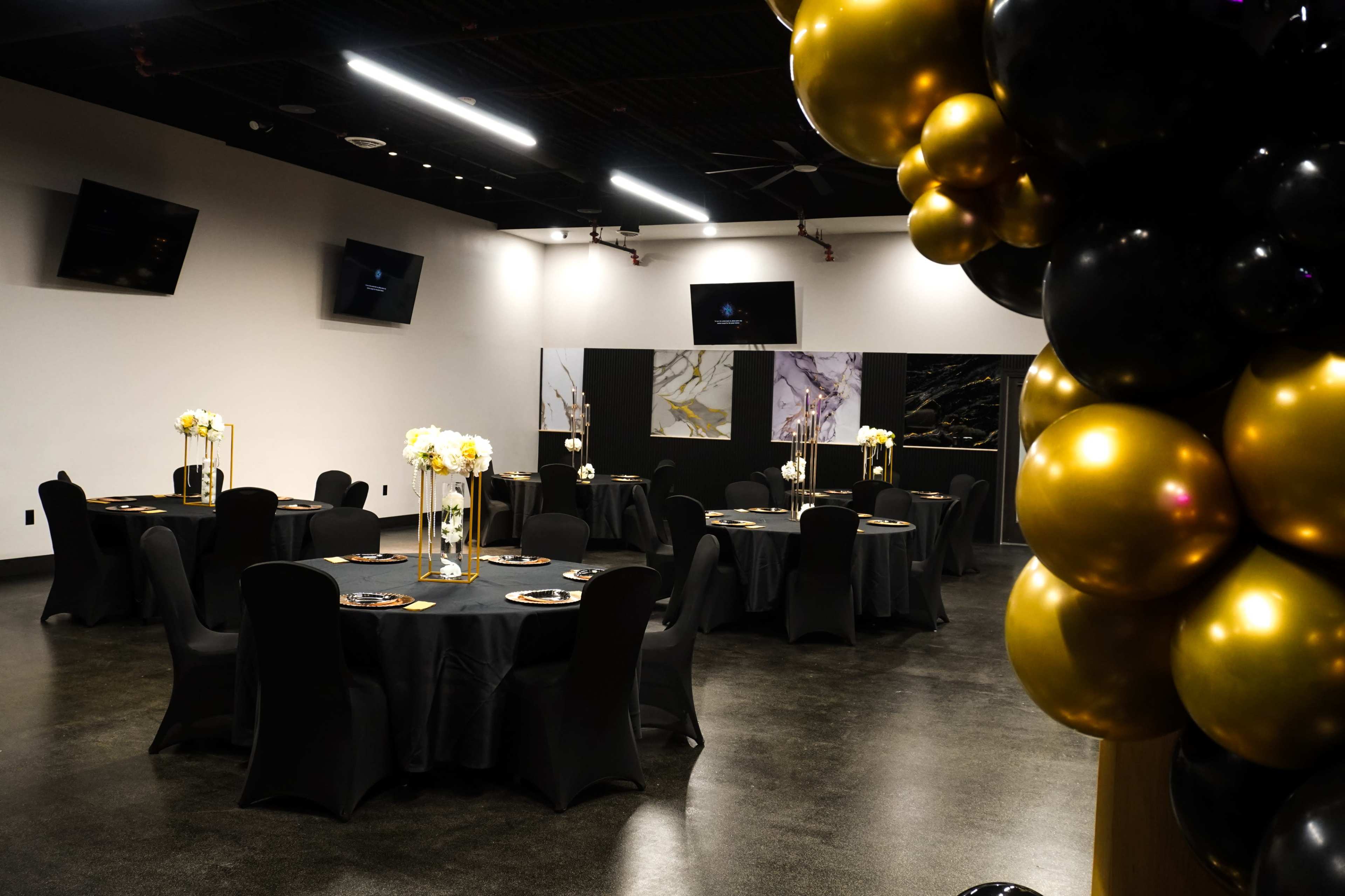 A banquet hall features several round tables covered with black tablecloths, adorned with floral centerpieces, and decorated with gold and black balloons.