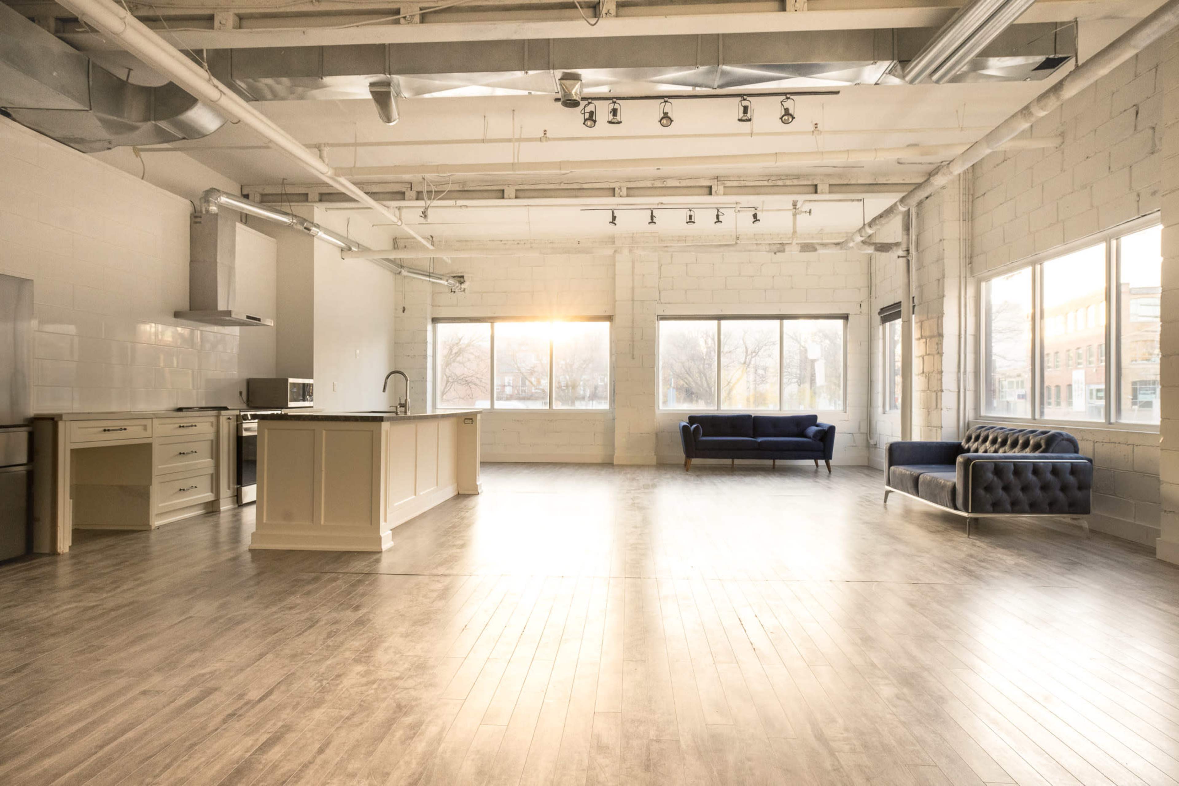 The image shows a spacious, minimalist interior with a modern kitchen on one side and a blue sofa on the opposite side, featuring large windows that allow natural light to enter.