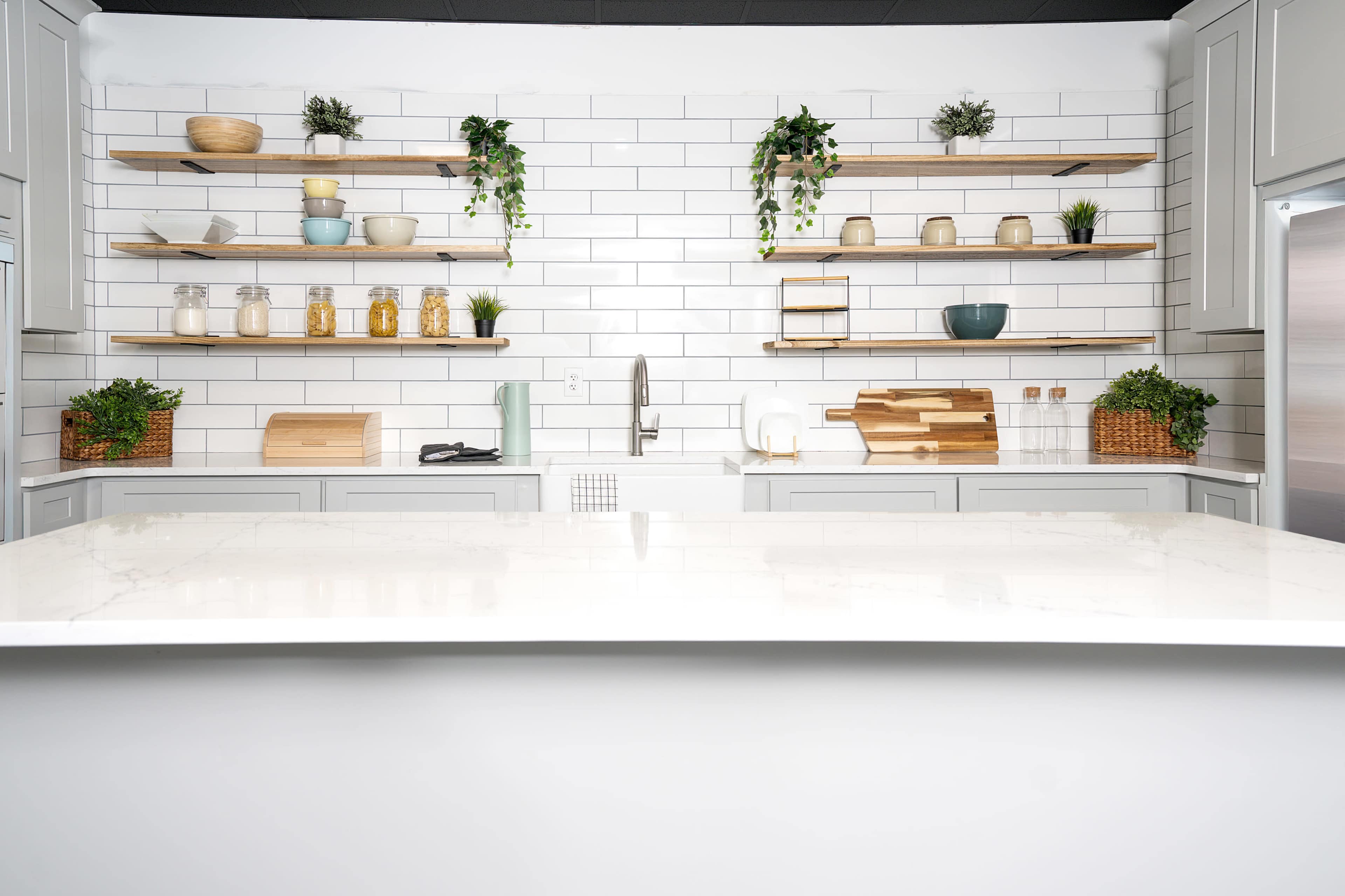 The kitchen features a white tiled wall with wooden shelves displaying decorative items and plants, alongside a sleek marble countertop.