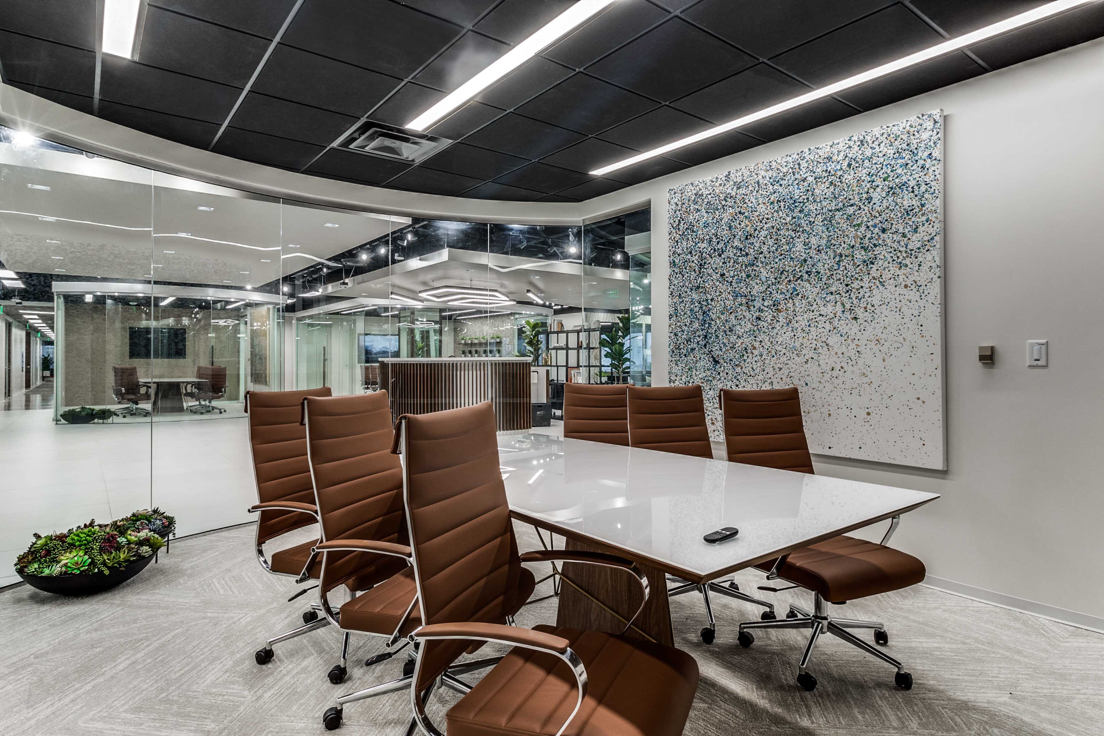 A modern conference room features a large glass table surrounded by six brown chairs, with a decorative wall art piece and glass walls separating the space from adjacent areas.