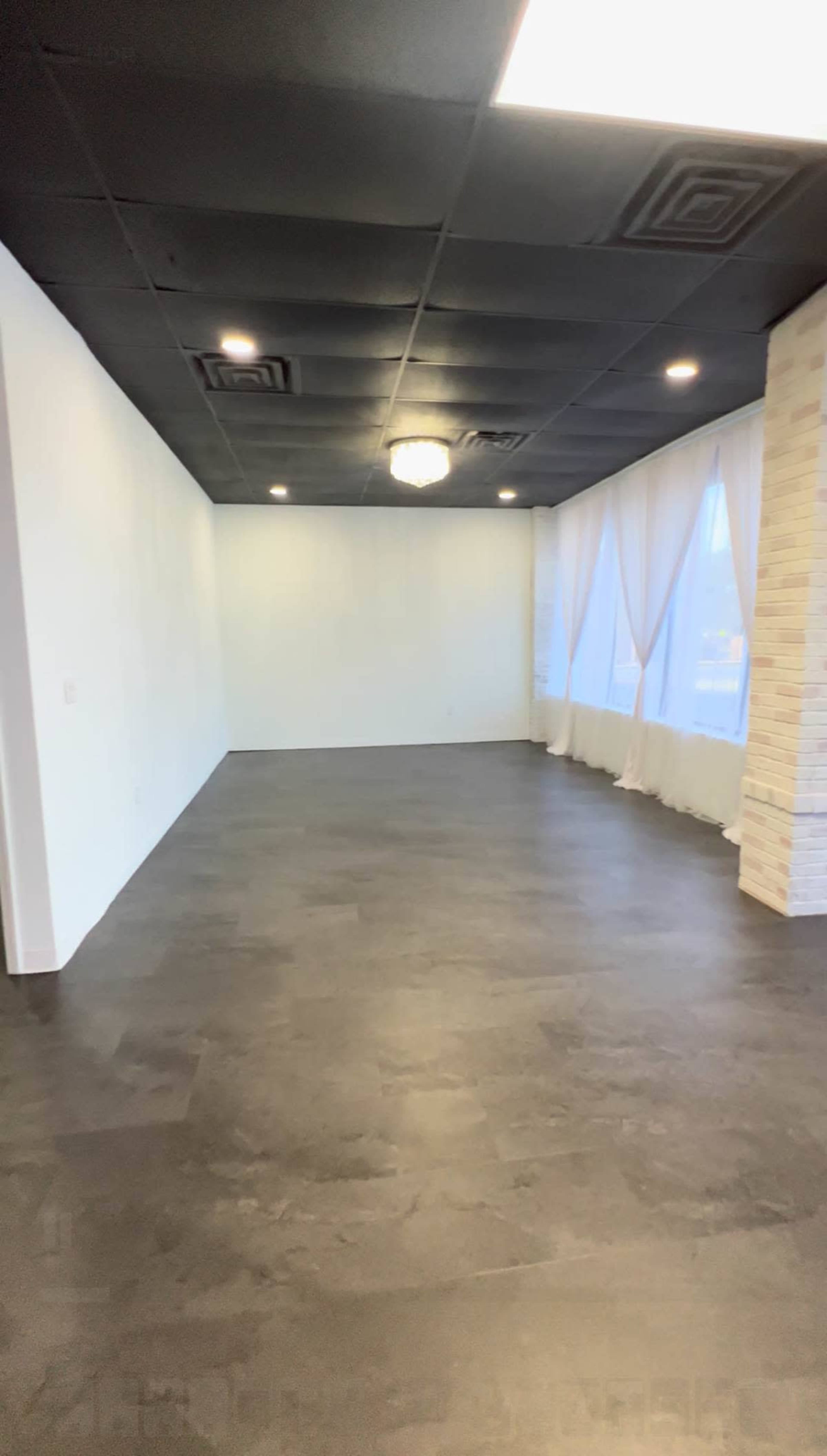 The image shows a spacious room with black flooring, white walls, and large windows draped with curtains.