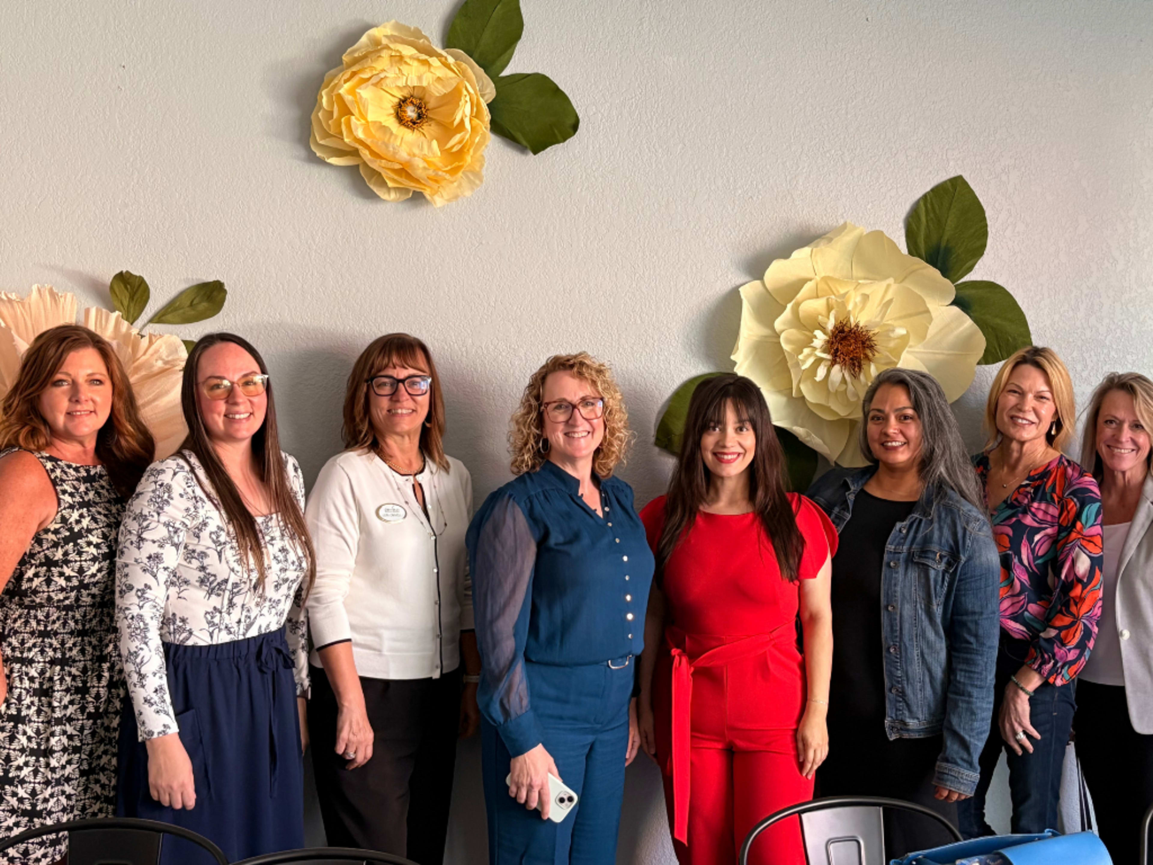 A group of nine women stands in front of large decorative flowers on a wall, all smiling for the camera in a well-lit setting.
