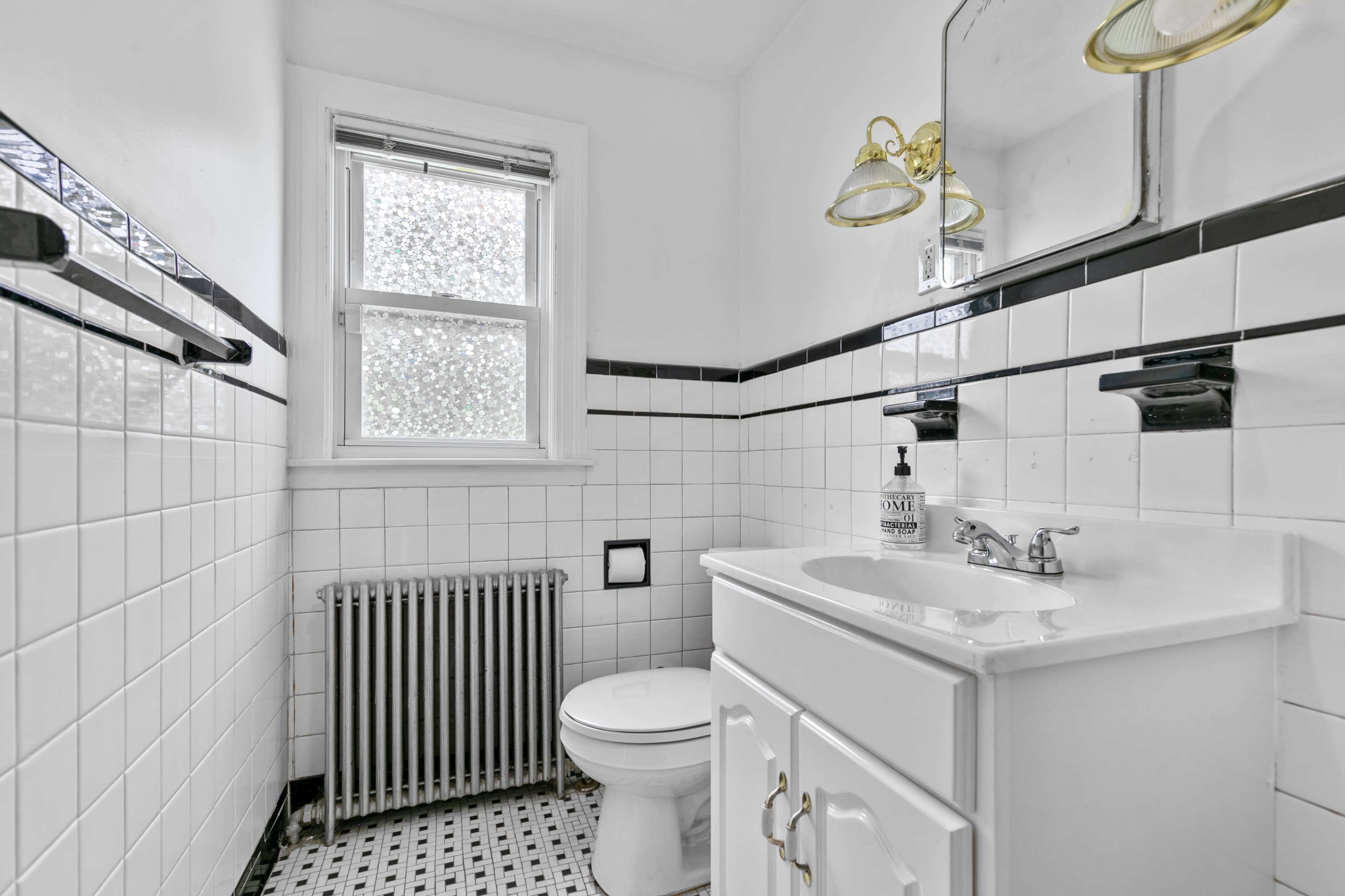 A small bathroom featuring white tiled walls, a black and white checkered floor, a white sink with a cabinet, a toilet, and a radiator.