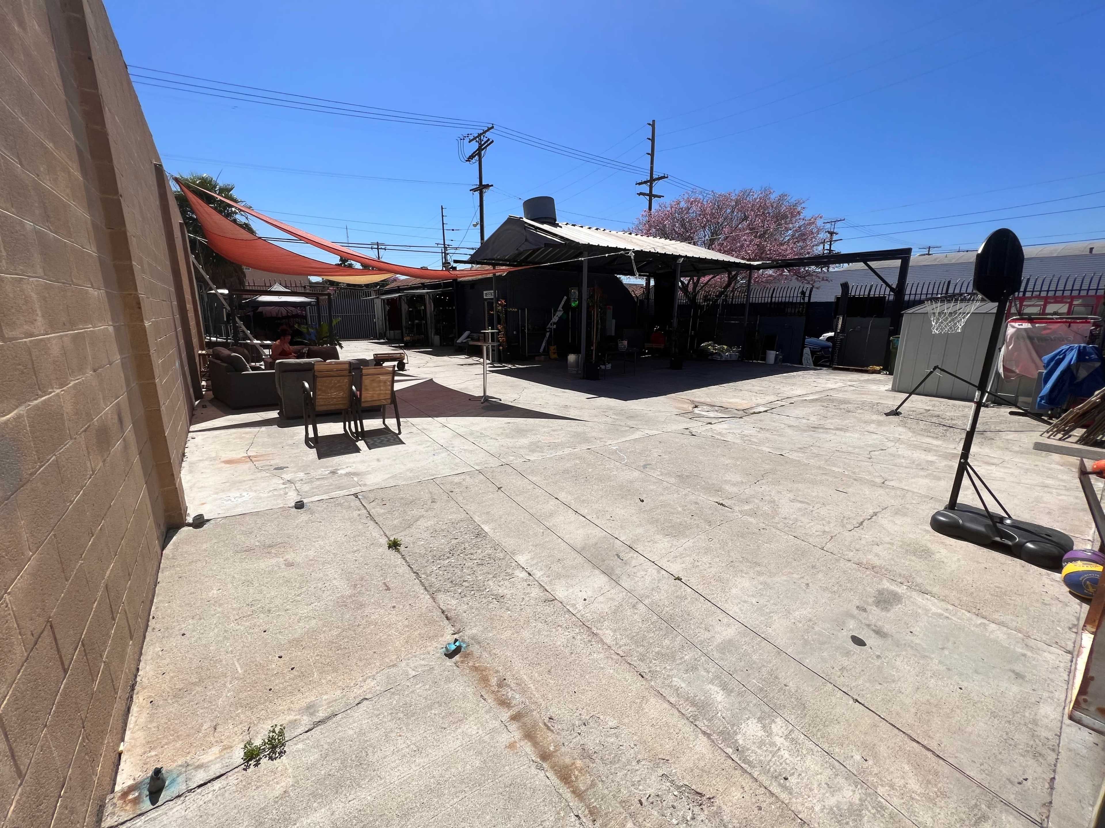 A large concrete outdoor space with various structures and furniture, including a covered area and a basketball hoop.