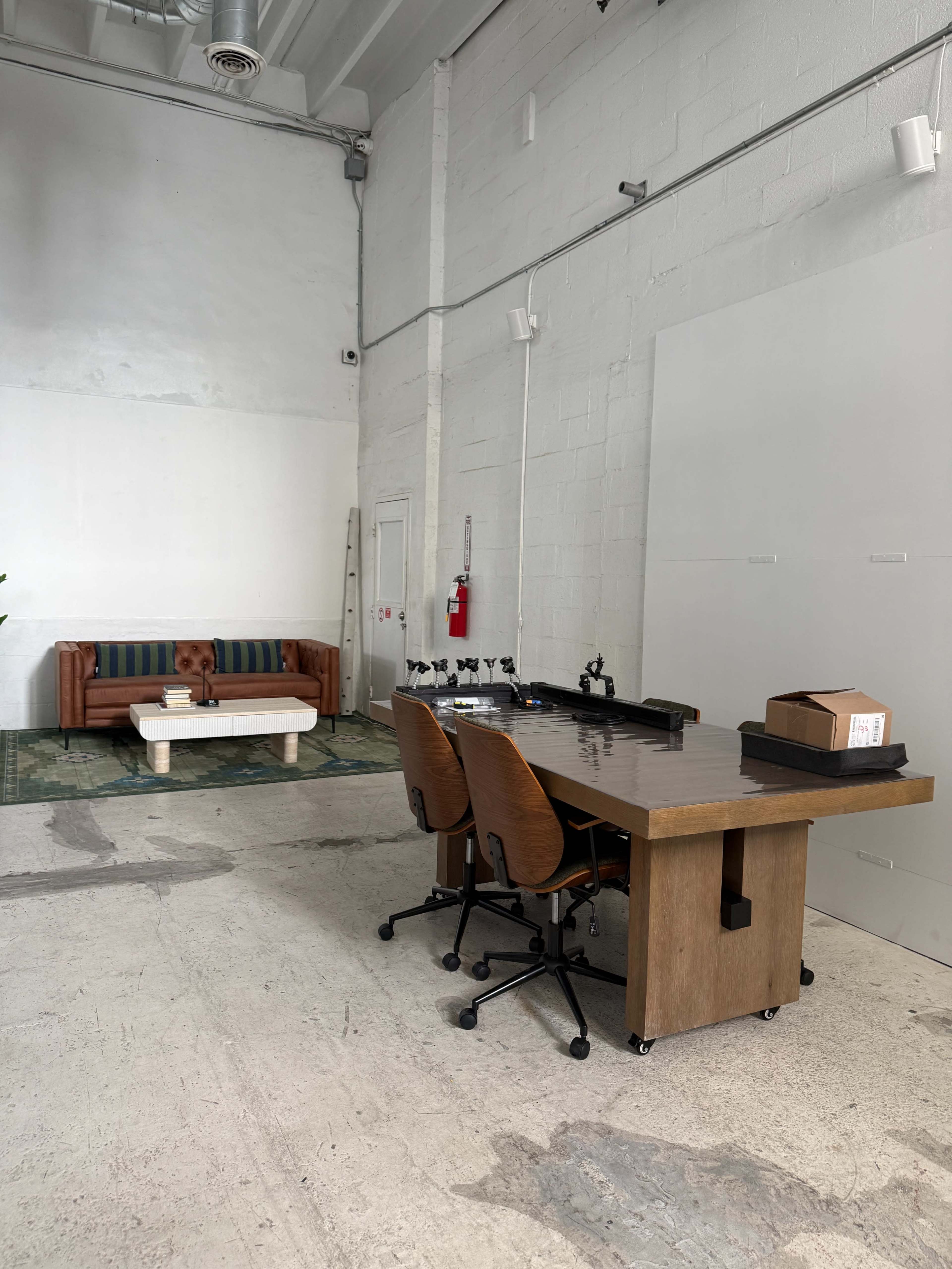 The image shows a spacious room featuring a wooden desk with two rolling chairs on one side and a brown sofa with a coffee table on the other side.