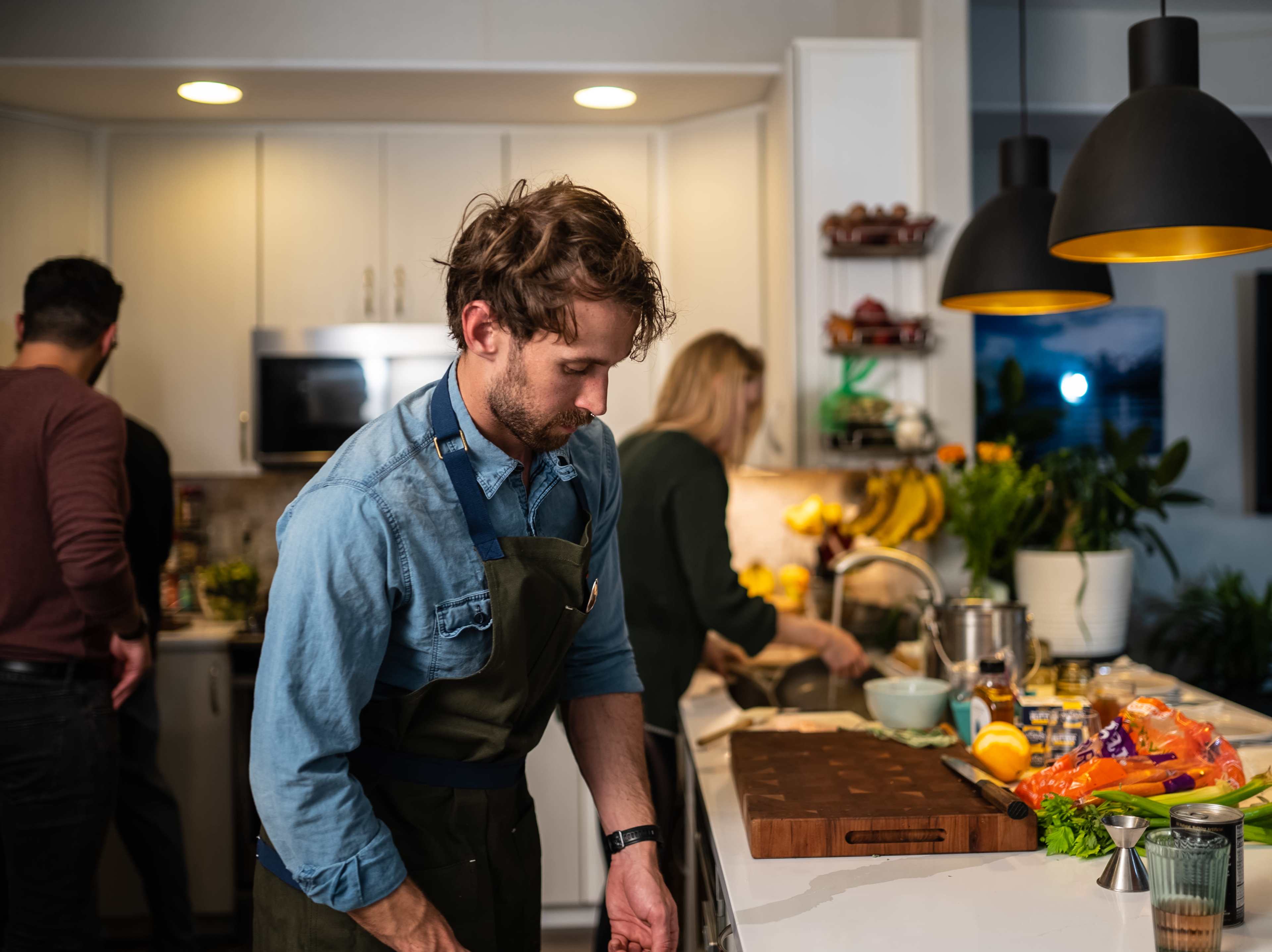 A man in an apron prepares food at a kitchen island while two other people work in the background.