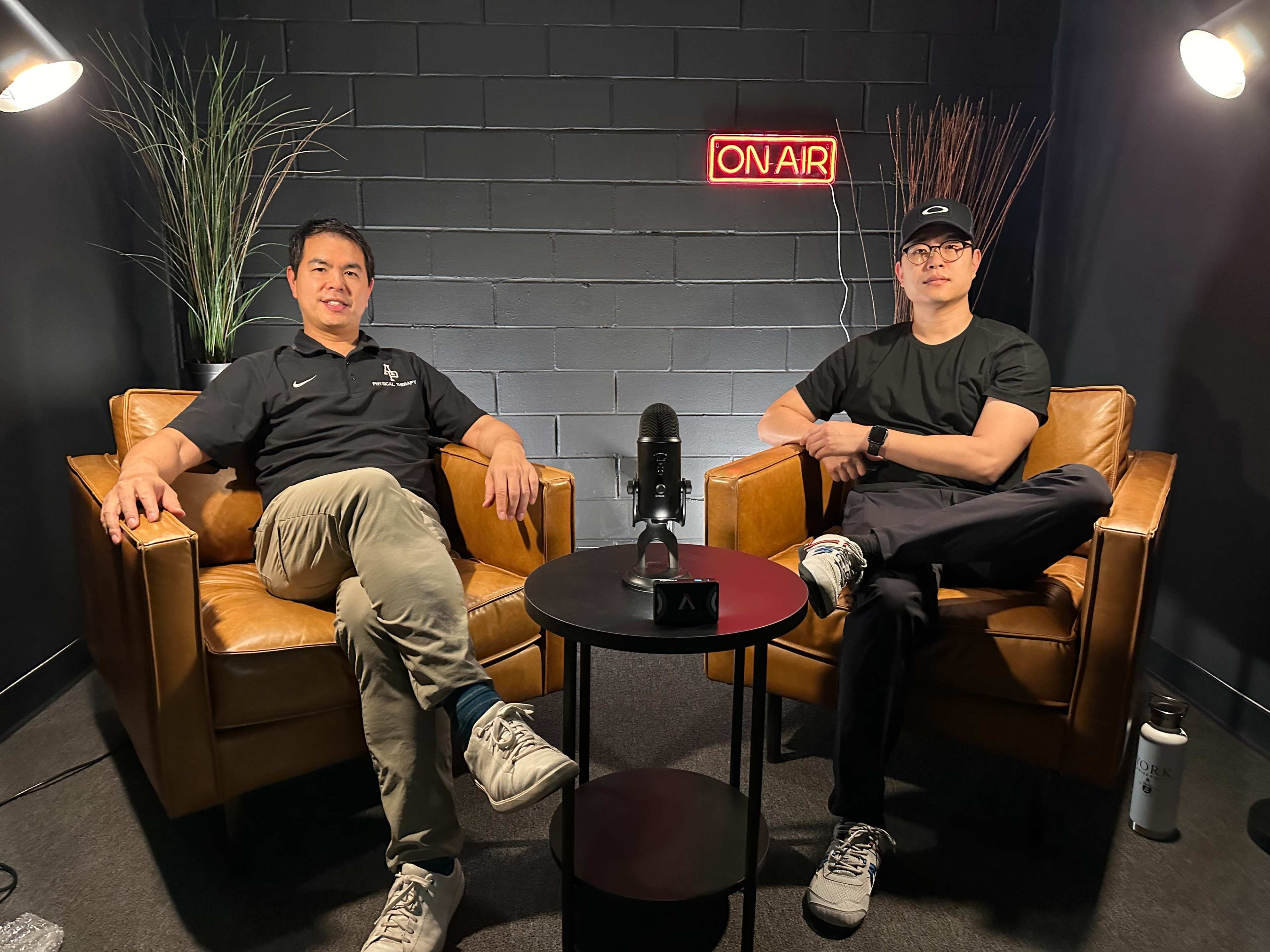 Two men sit in brown leather chairs in a studio setting, with an "ON AIR" sign illuminated in the background and a microphone on a table between them.