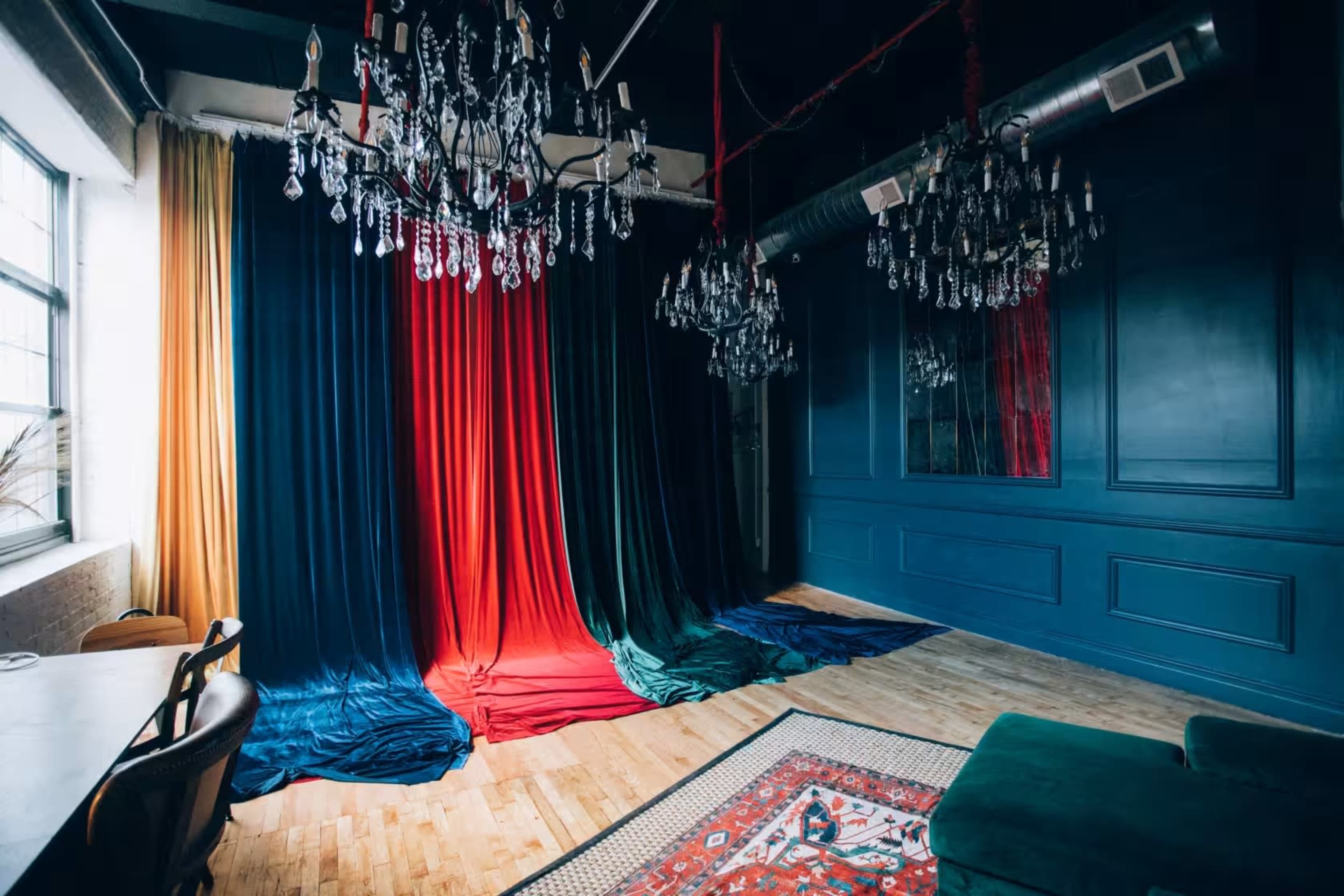The image shows a stylish interior space featuring large velvet curtains in shades of red, green, and blue, complemented by ornate chandeliers and a patterned rug on the wooden floor.