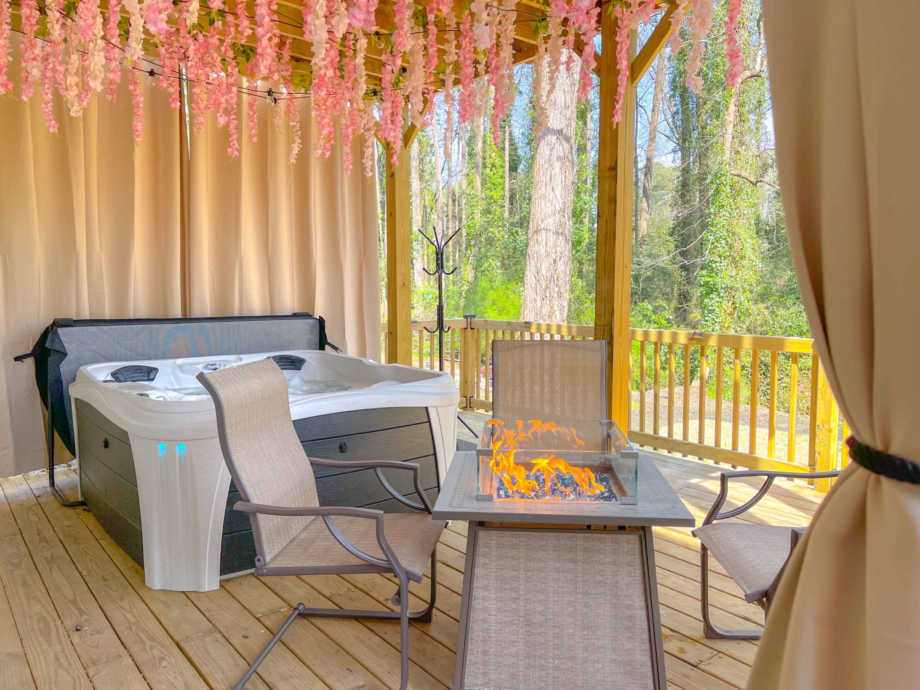 The scene features a hot tub under a canopy decorated with hanging flowers, accompanied by a fire table and several chairs on a wooden deck.