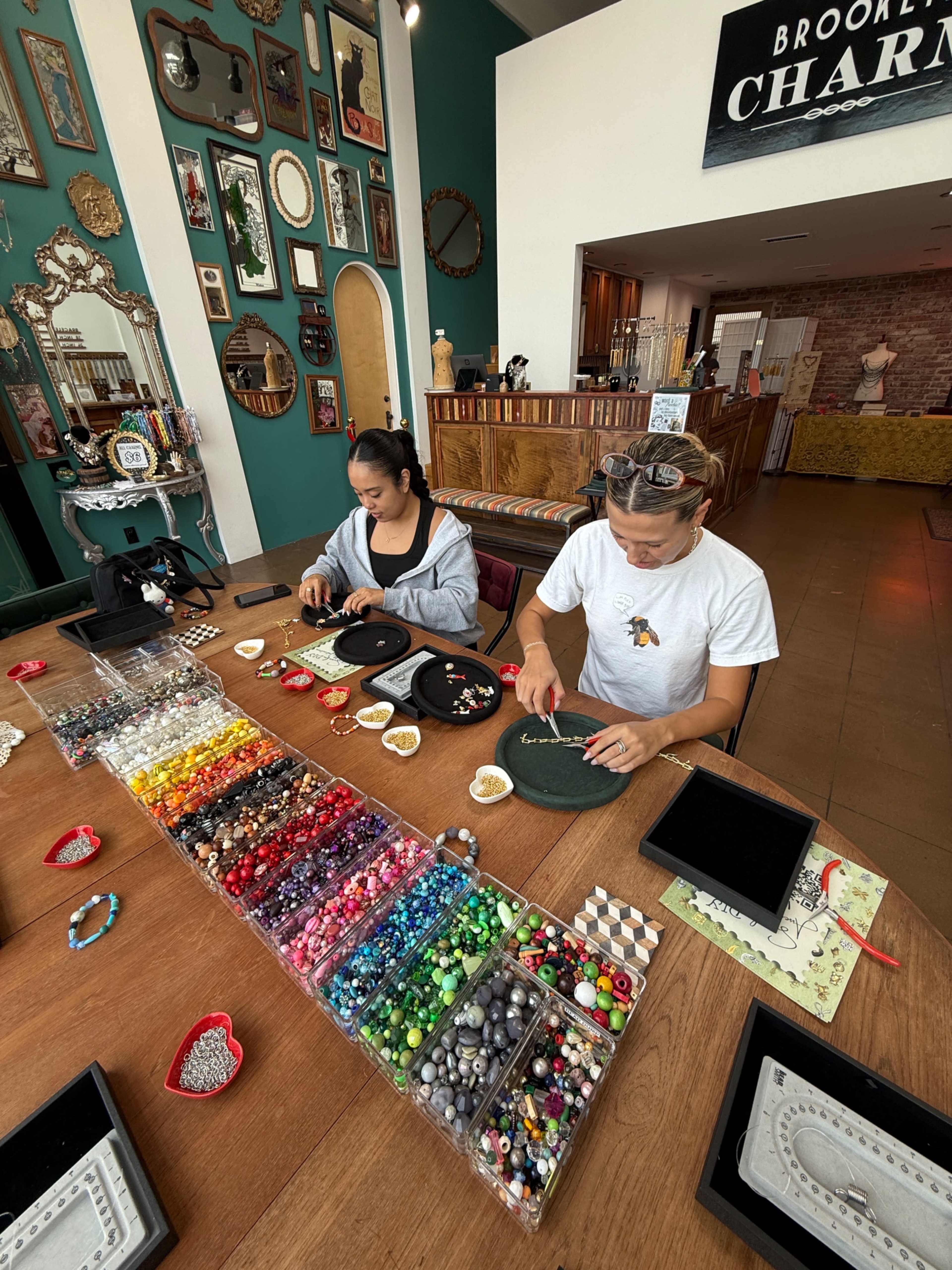 Two individuals are focused on crafting with various colorful beads laid out on a table in a shop.