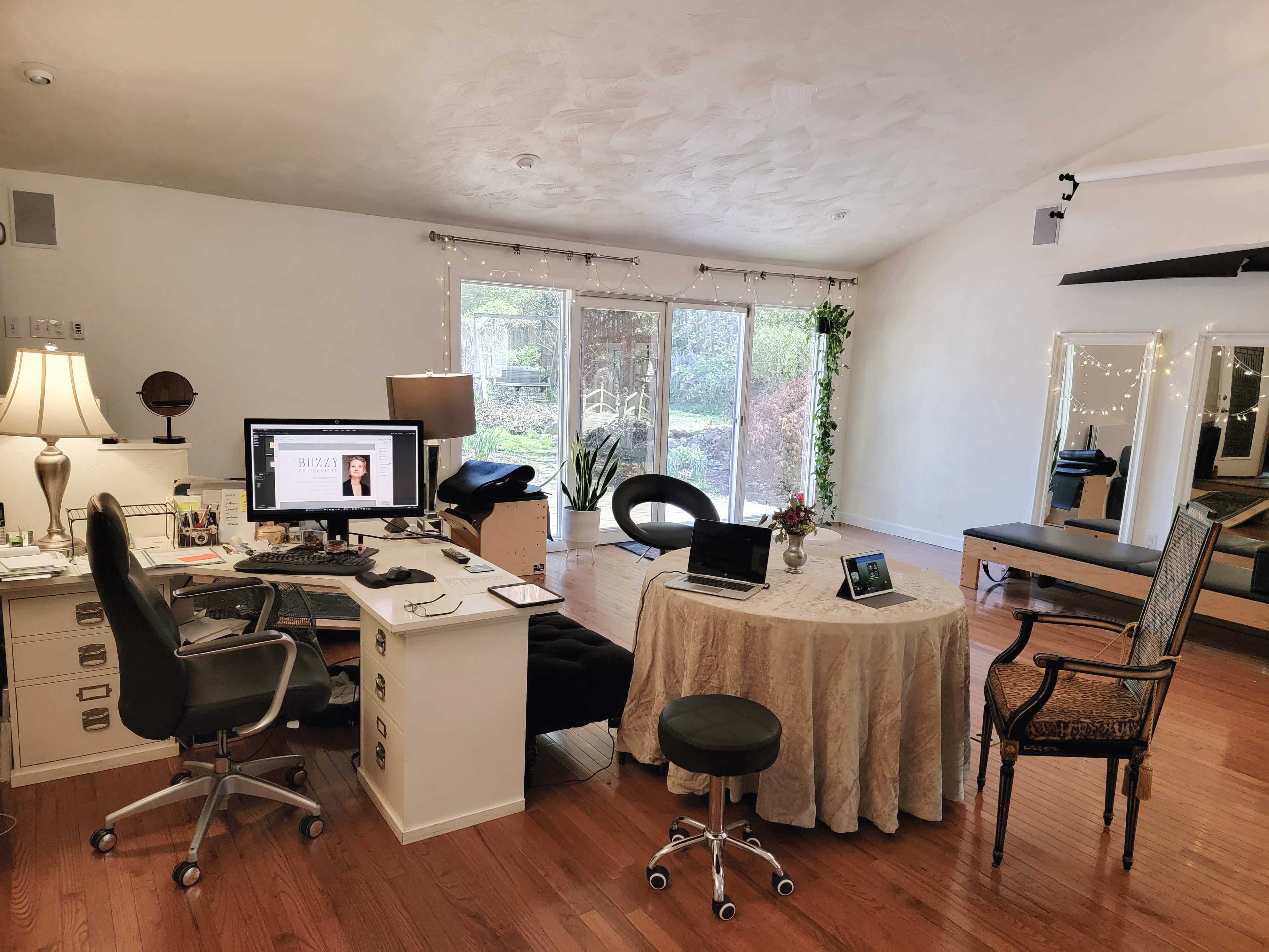The image shows a well-lit room featuring a desk with a computer, a table with a tablecloth, a chair, and a large mirror along with large windows that overlook a garden.