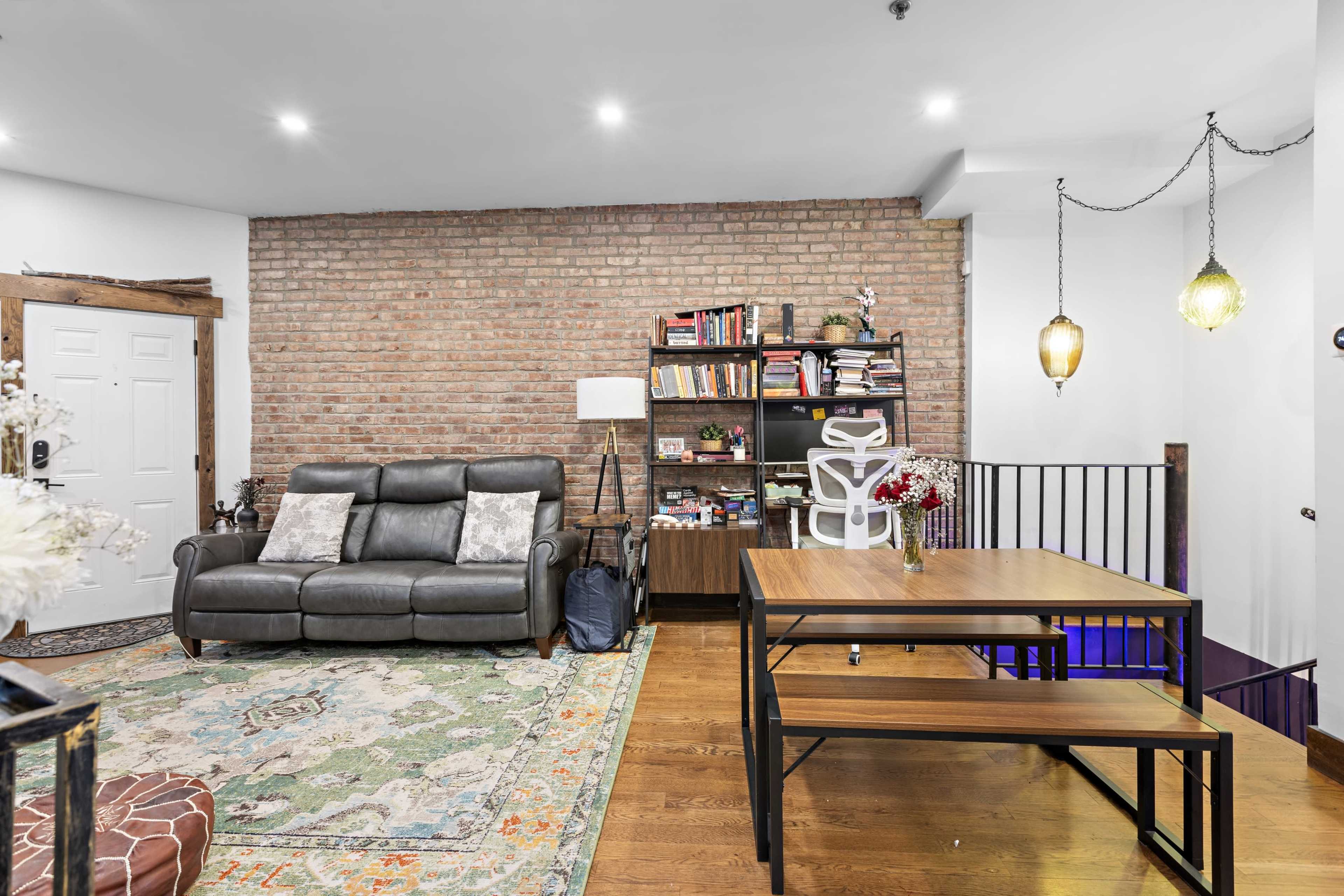 The scene shows a cozy living area with a leather sofa, a wooden table, bookshelves filled with books, and exposed brick walls.