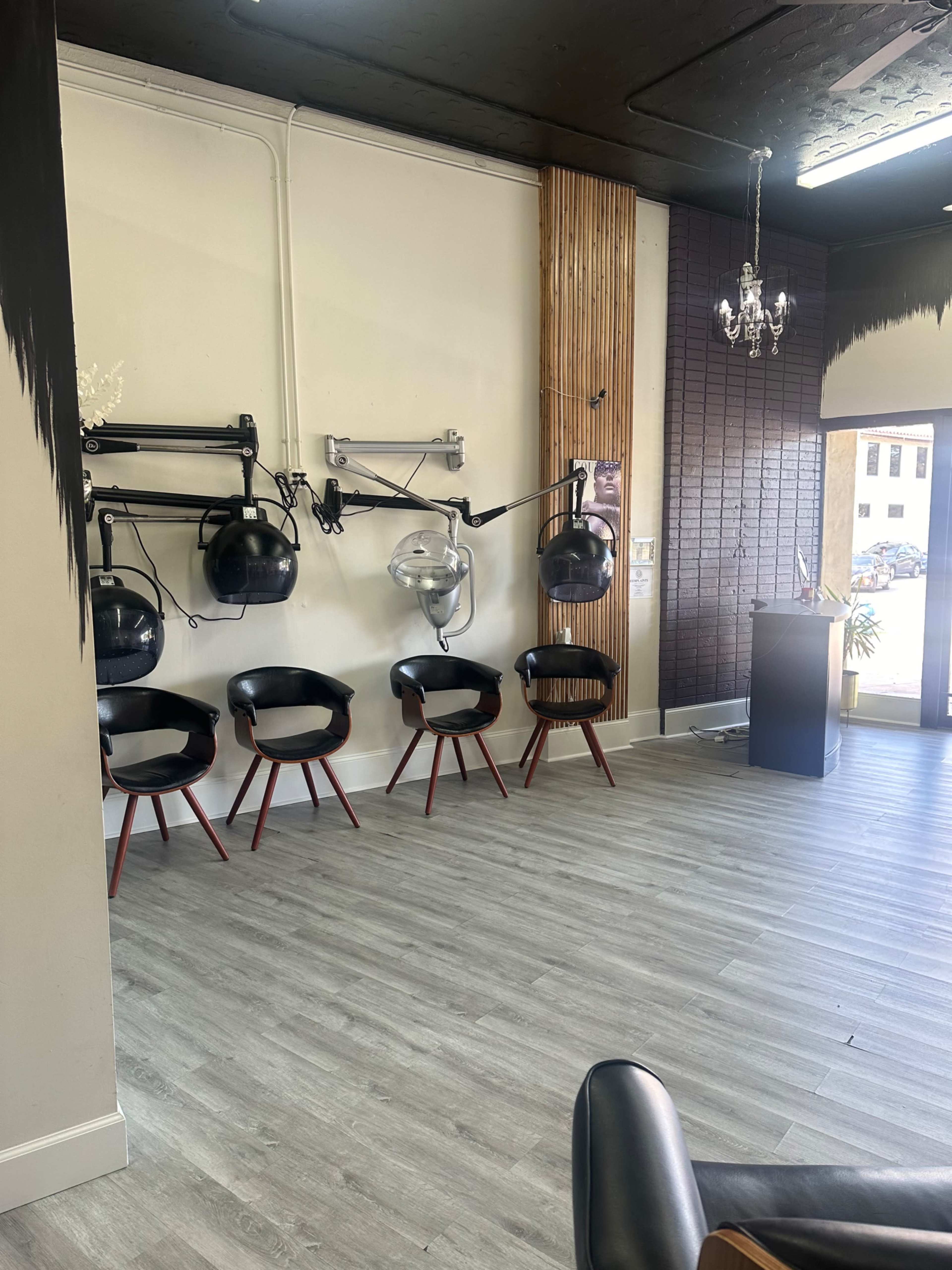 A modern hair salon interior with several black chairs arranged in front of wall-mounted hairdryers and a reception area partially visible in the background.