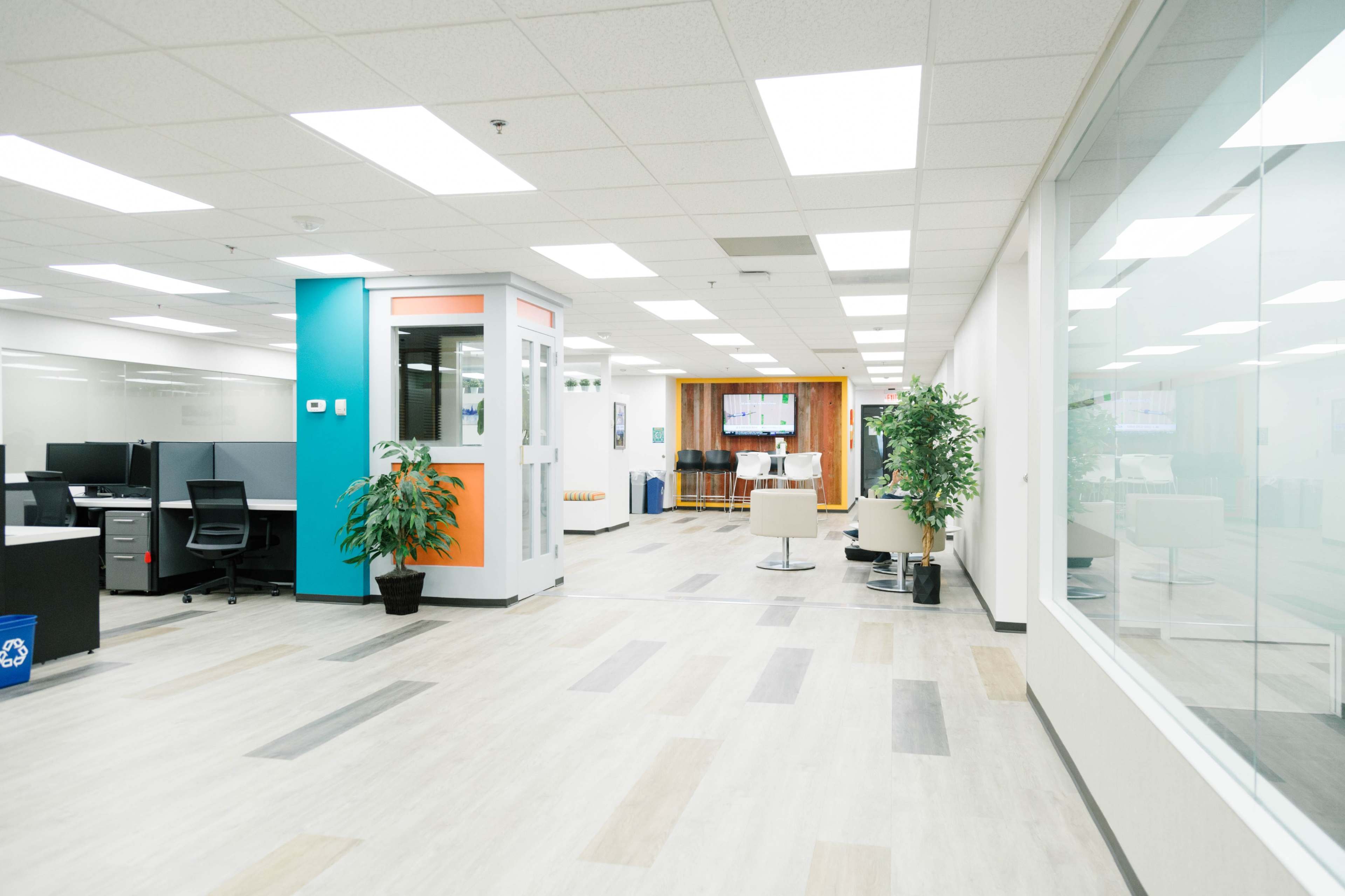 A modern office corridor featuring shared workspaces, a glass-walled meeting room, and plants arranged throughout.