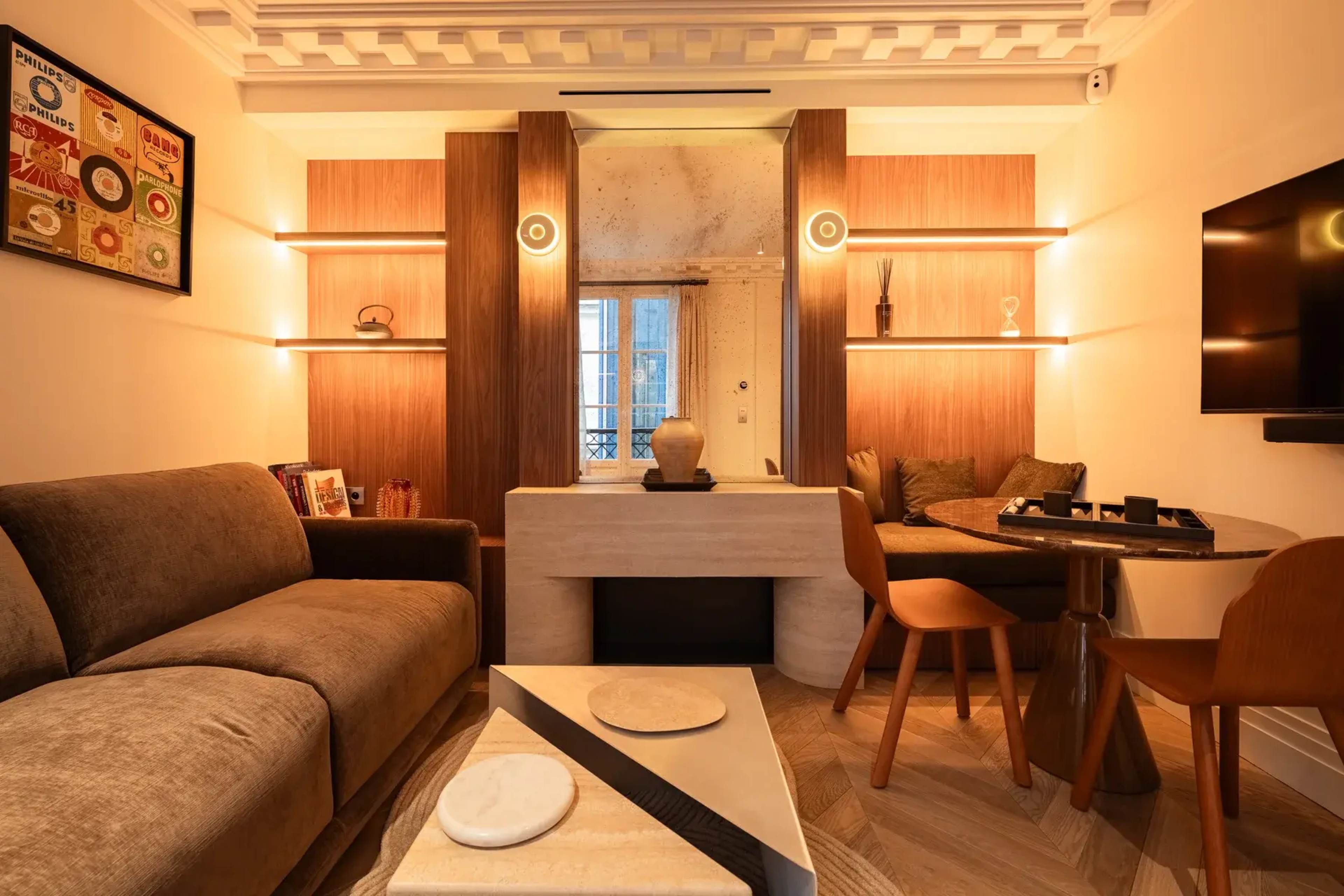 The image shows a modern living space with a sofa, a small dining table, and decorative elements, featuring a neutral color palette and contemporary furnishings.