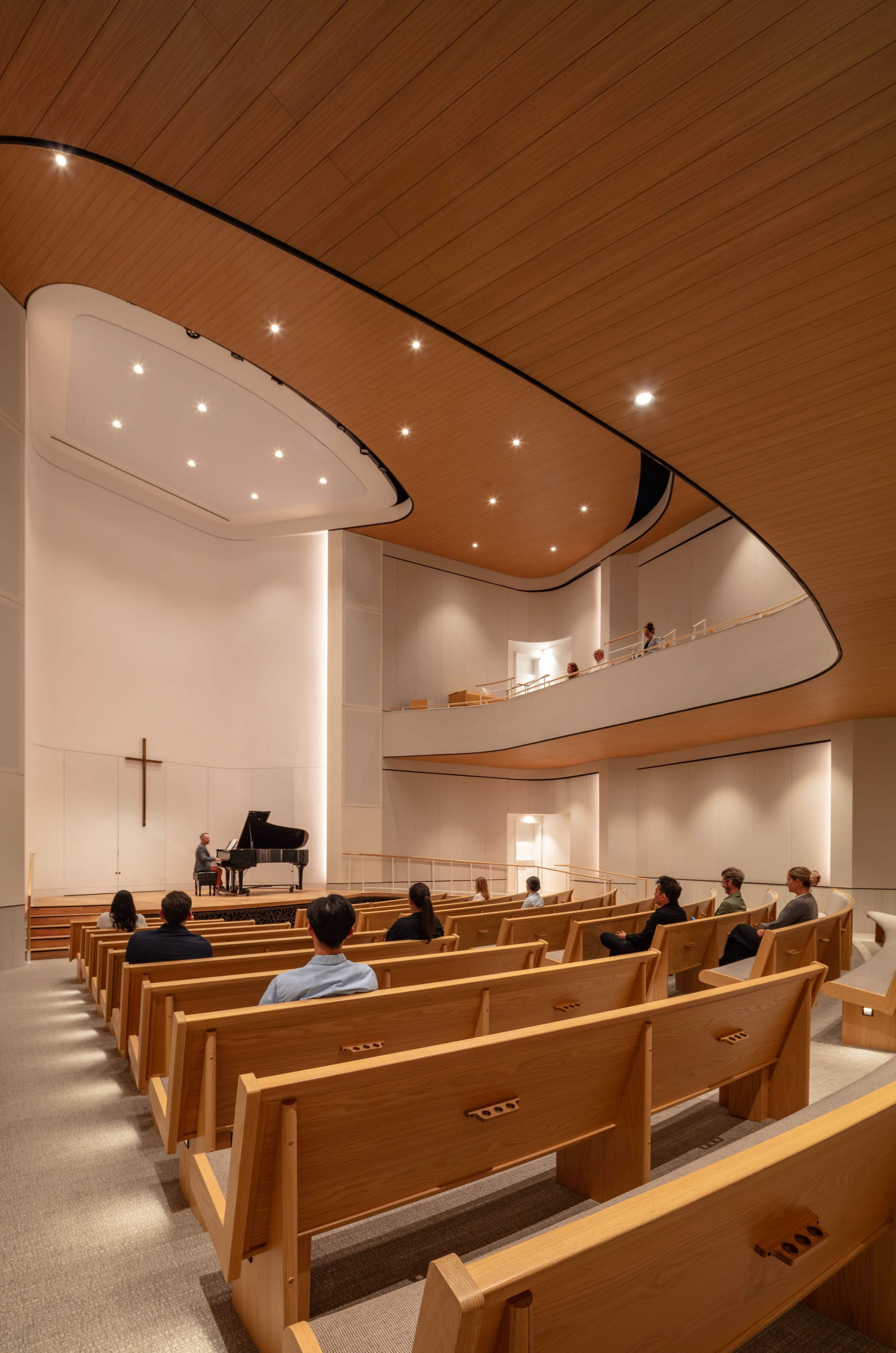 A modern church interior features wooden pews facing a grand piano on a stage, with a cross on the wall and an upper balcony visible.