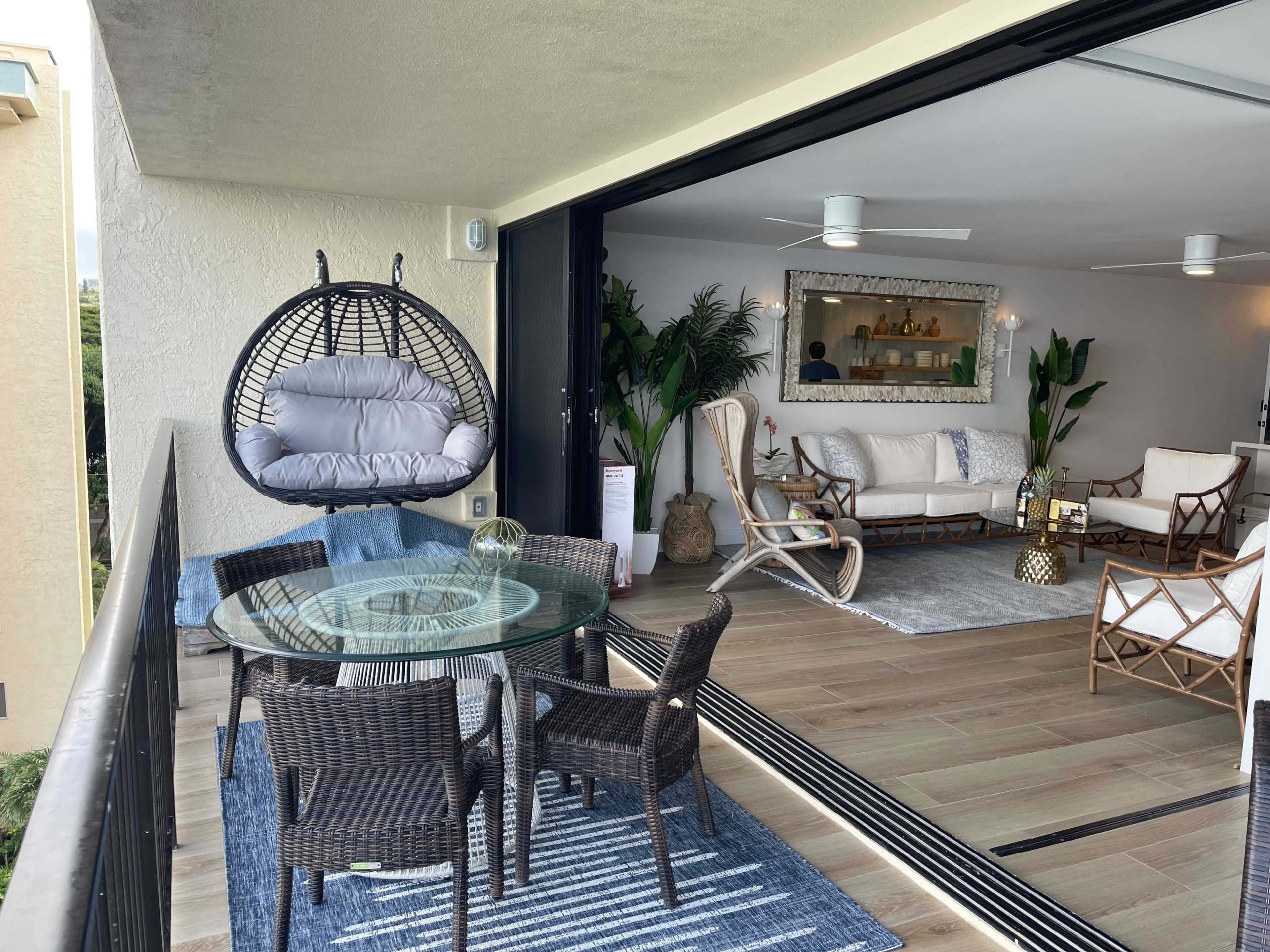A spacious balcony with a glass dining table and wicker chairs, overlooking a modern living area furnished with plants and decorative elements.
