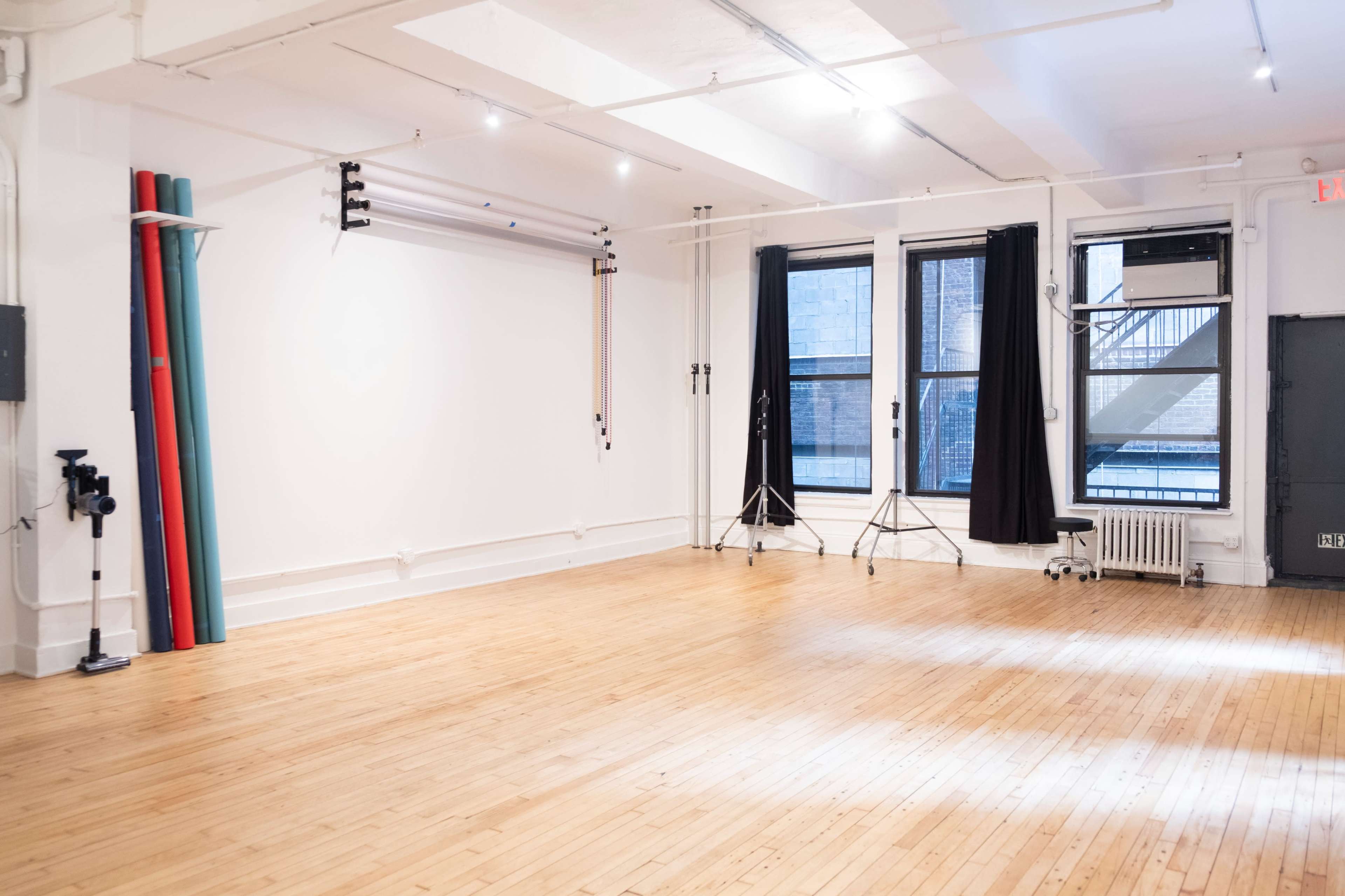 The image shows a spacious, well-lit studio with wooden floors, a white wall, and equipment such as light stands and colored backdrops leaning against the wall.
