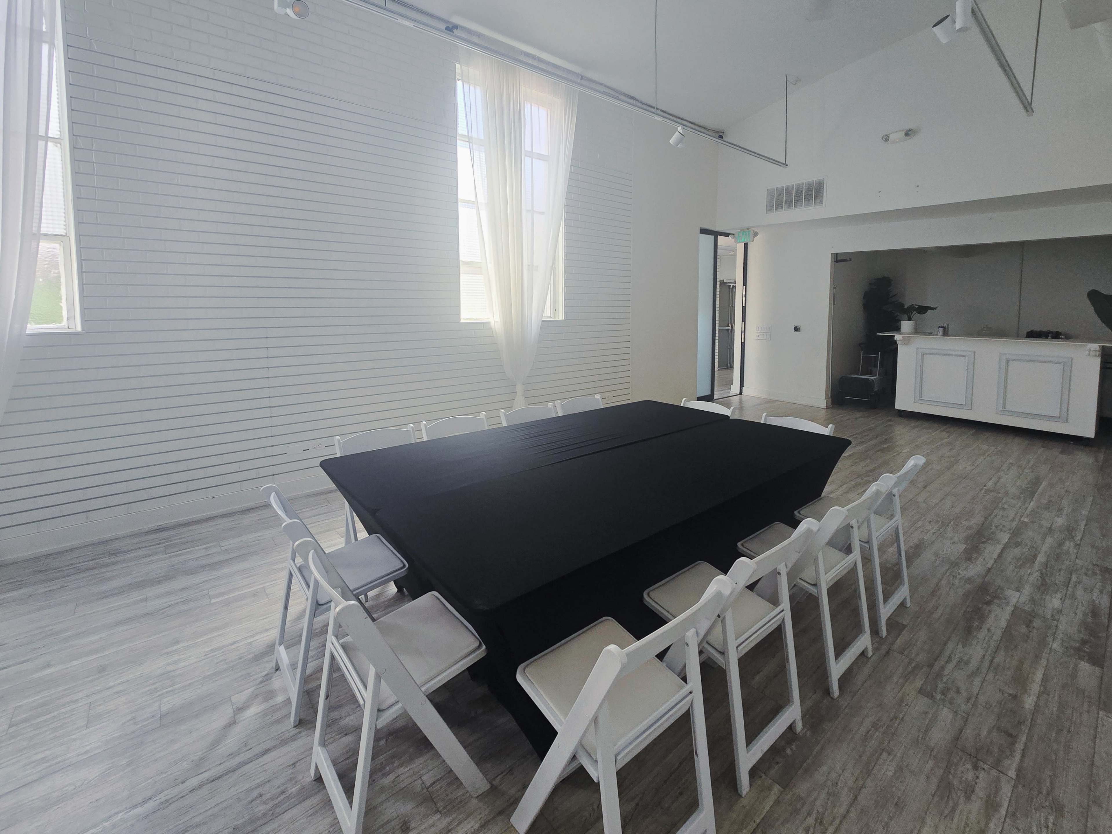 A rectangular table covered with a black tablecloth is surrounded by white folding chairs in a bright, airy event space with large windows.