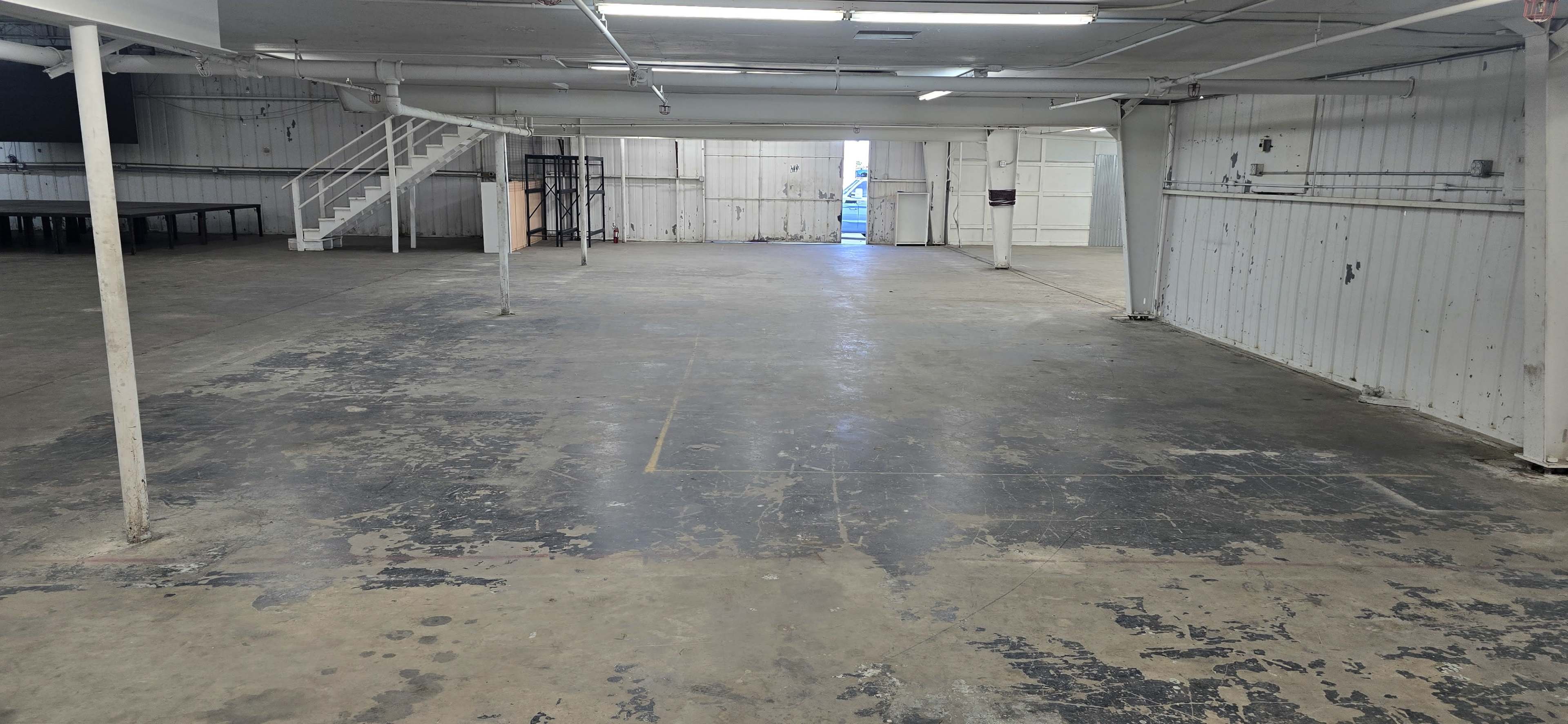 The image shows an empty, industrial-style interior space with concrete floors, white walls, and a set of stairs in the background leading to a higher level.