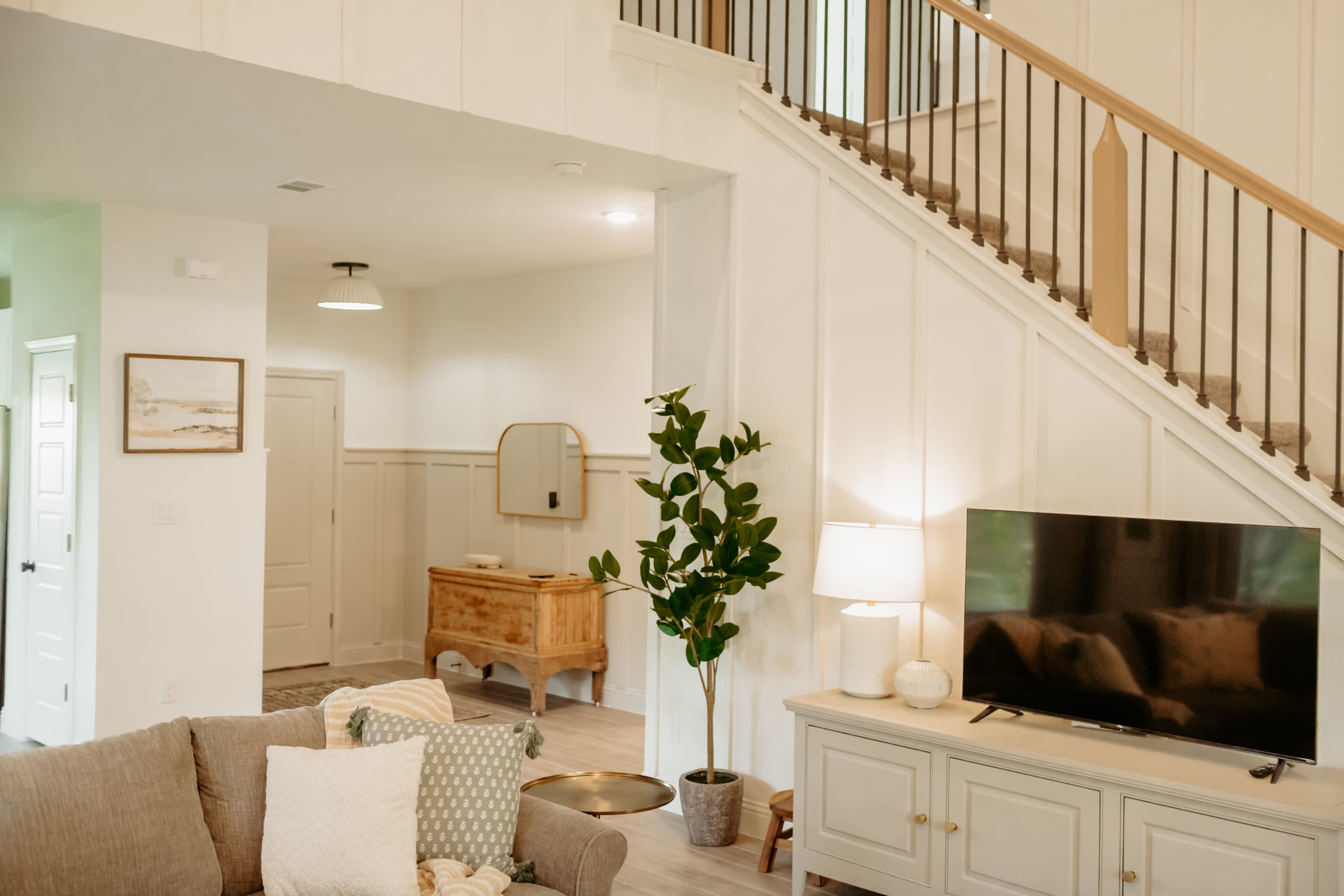 The image shows a bright and modern living space featuring a staircase, a television on a cabinet, a potted plant, and a small entryway with a mirror and a wooden chest.