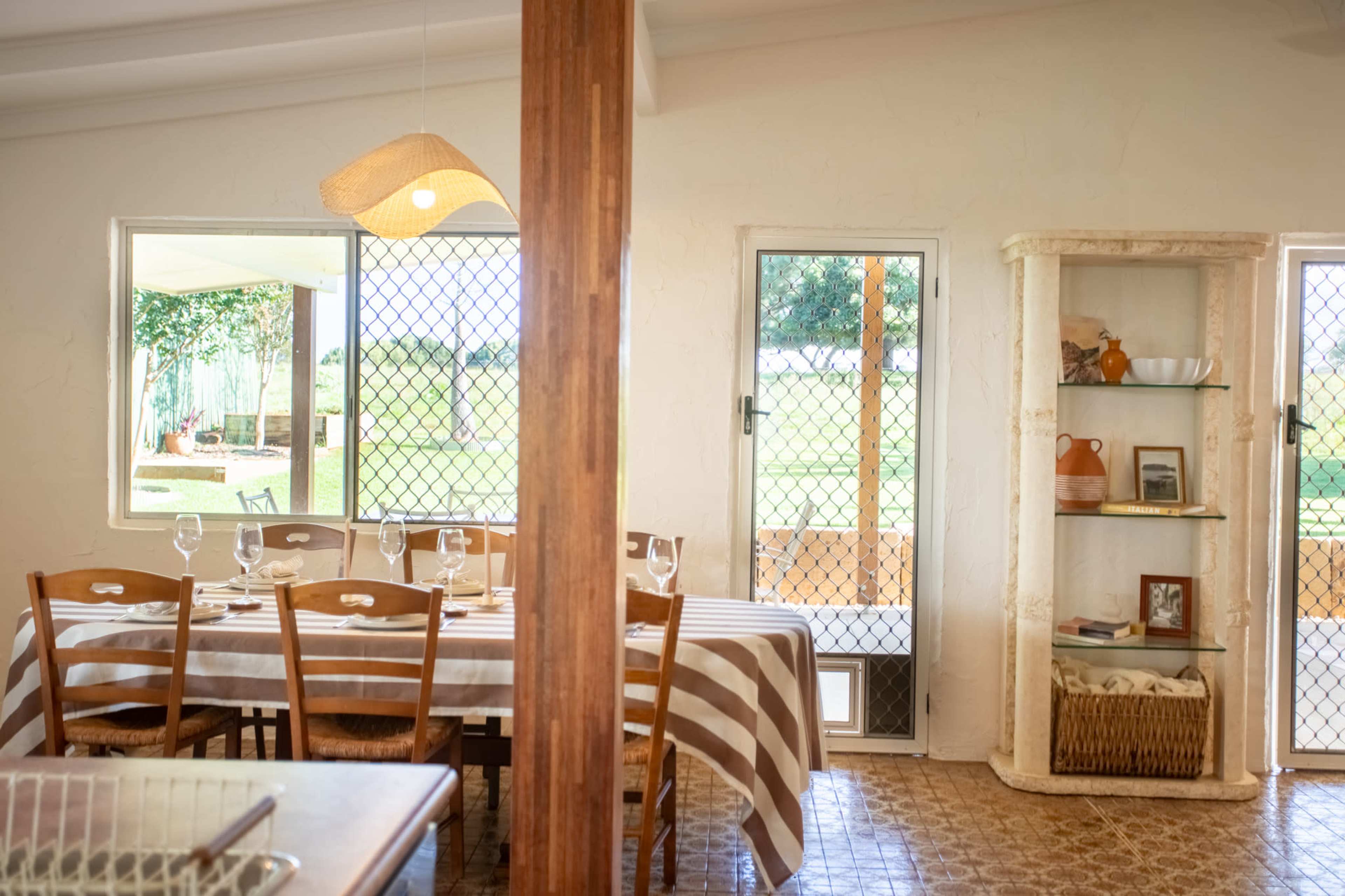 The image shows a dining area with a table set with striped tablecloths and chairs, adjacent to a shelf displaying various items and large windows overlooking a green outdoor space.