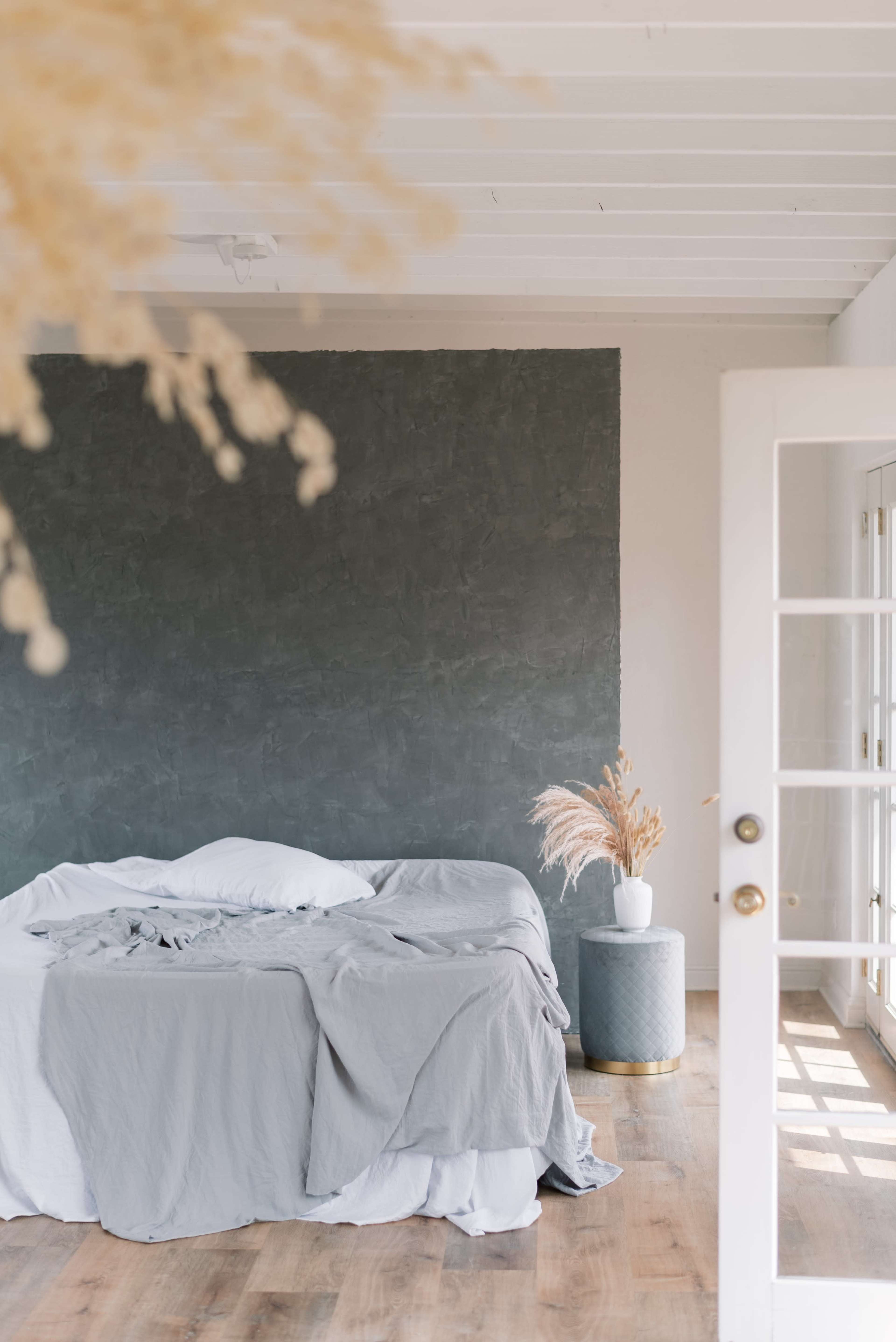 A bedroom features a bed with unmade gray linens and a decorative vase with dried plants beside a glass-paned door leading to an outdoor area.
