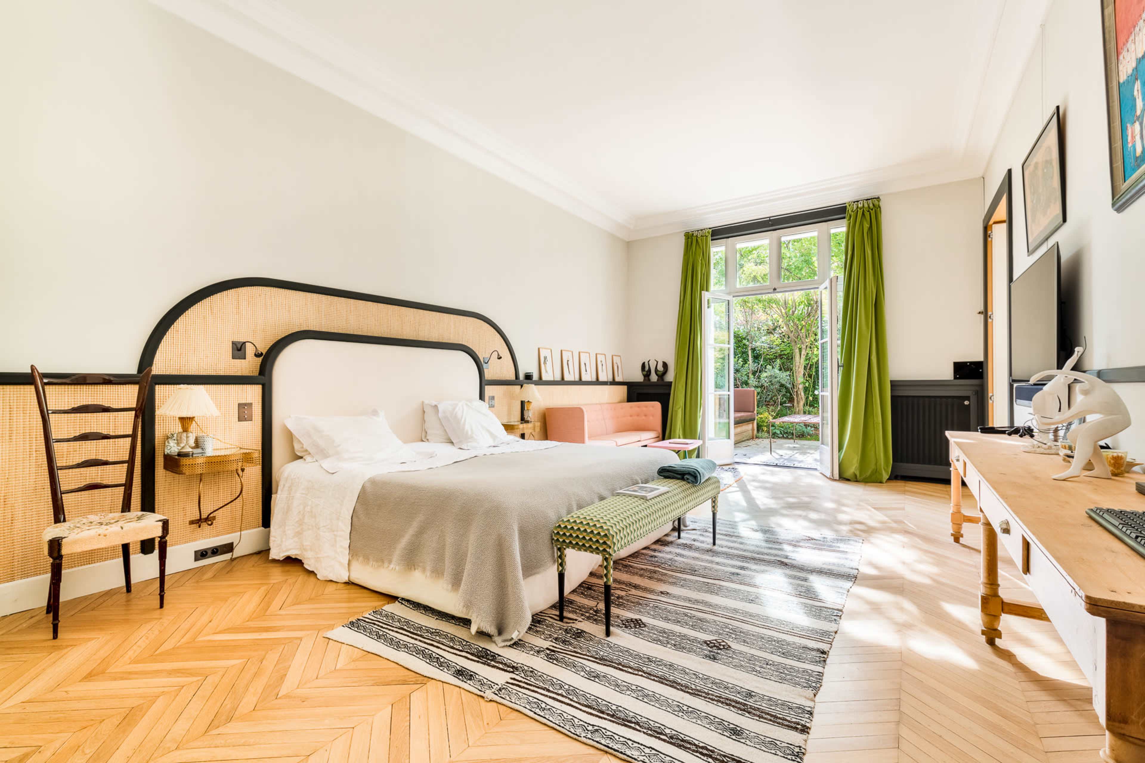 The image shows a bright bedroom featuring a large bed, a decorative chair, a wooden table, and a glass door leading to a garden area.