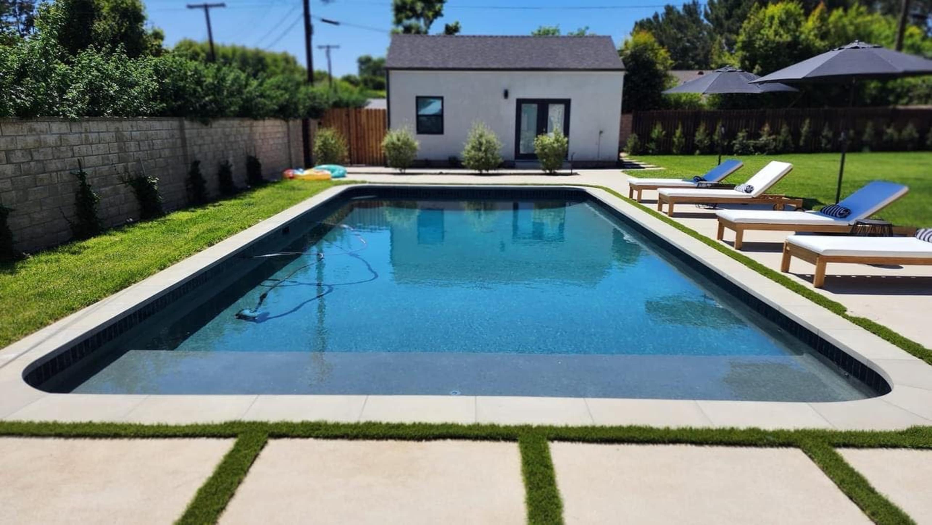 A clear swimming pool is surrounded by lounge chairs on a concrete patio, with a garden area and a small building in the background.