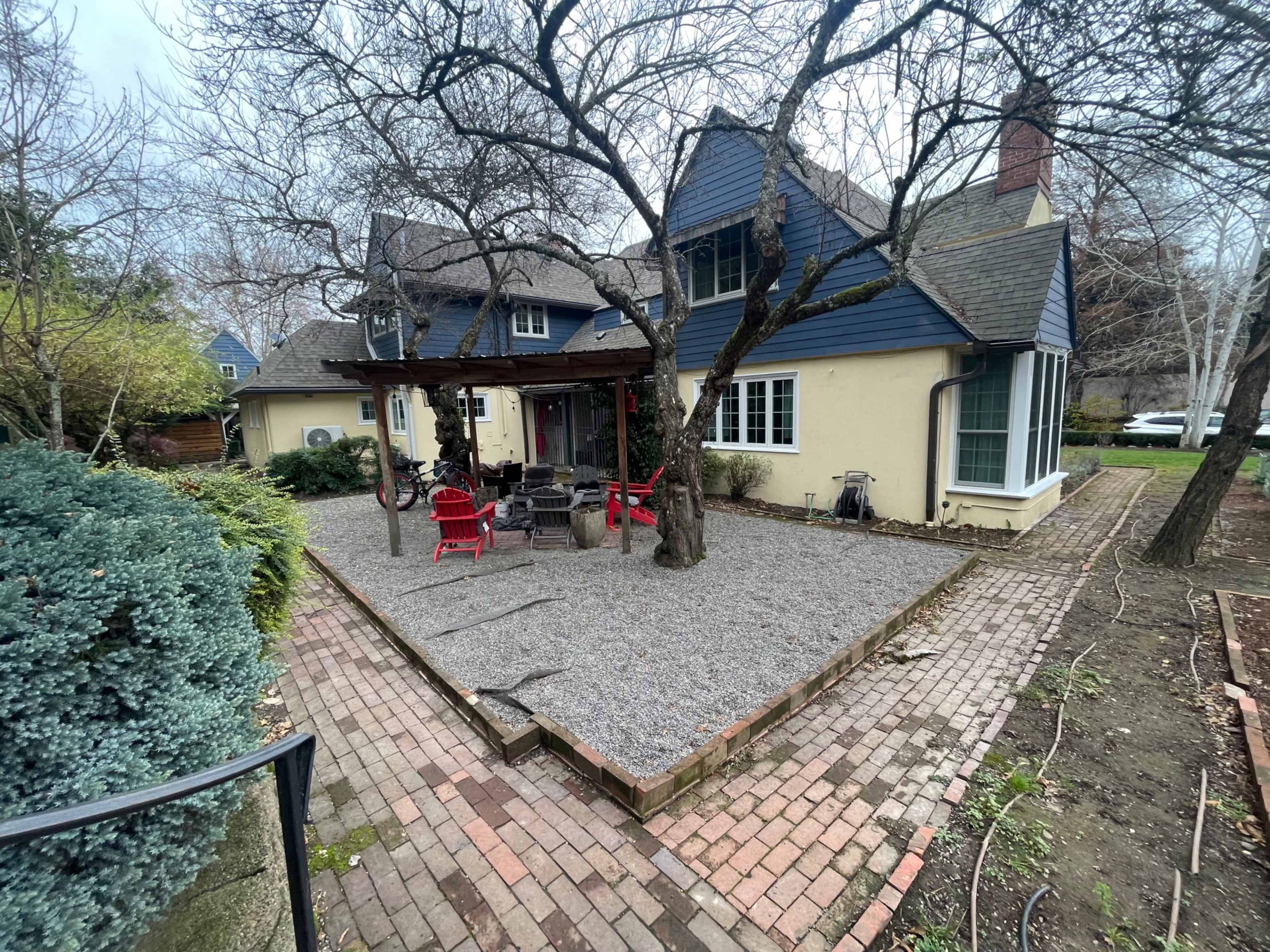 A backyard with a gravel area surrounded by trees, a patio with red chairs, and a house featuring a mix of blue and beige siding.