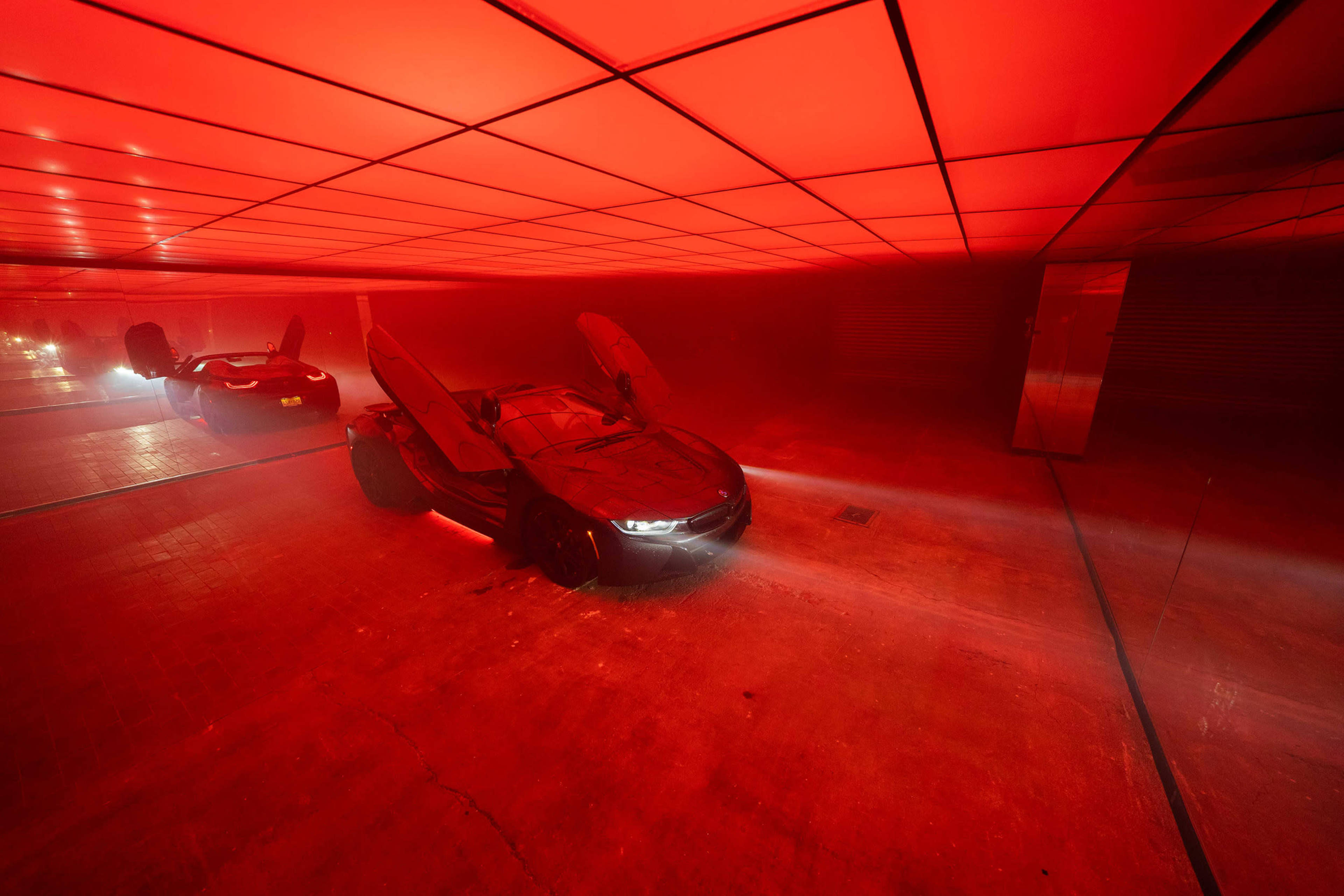 A sleek black sports car with its doors opened is positioned in a dimly lit garage illuminated by red lights.