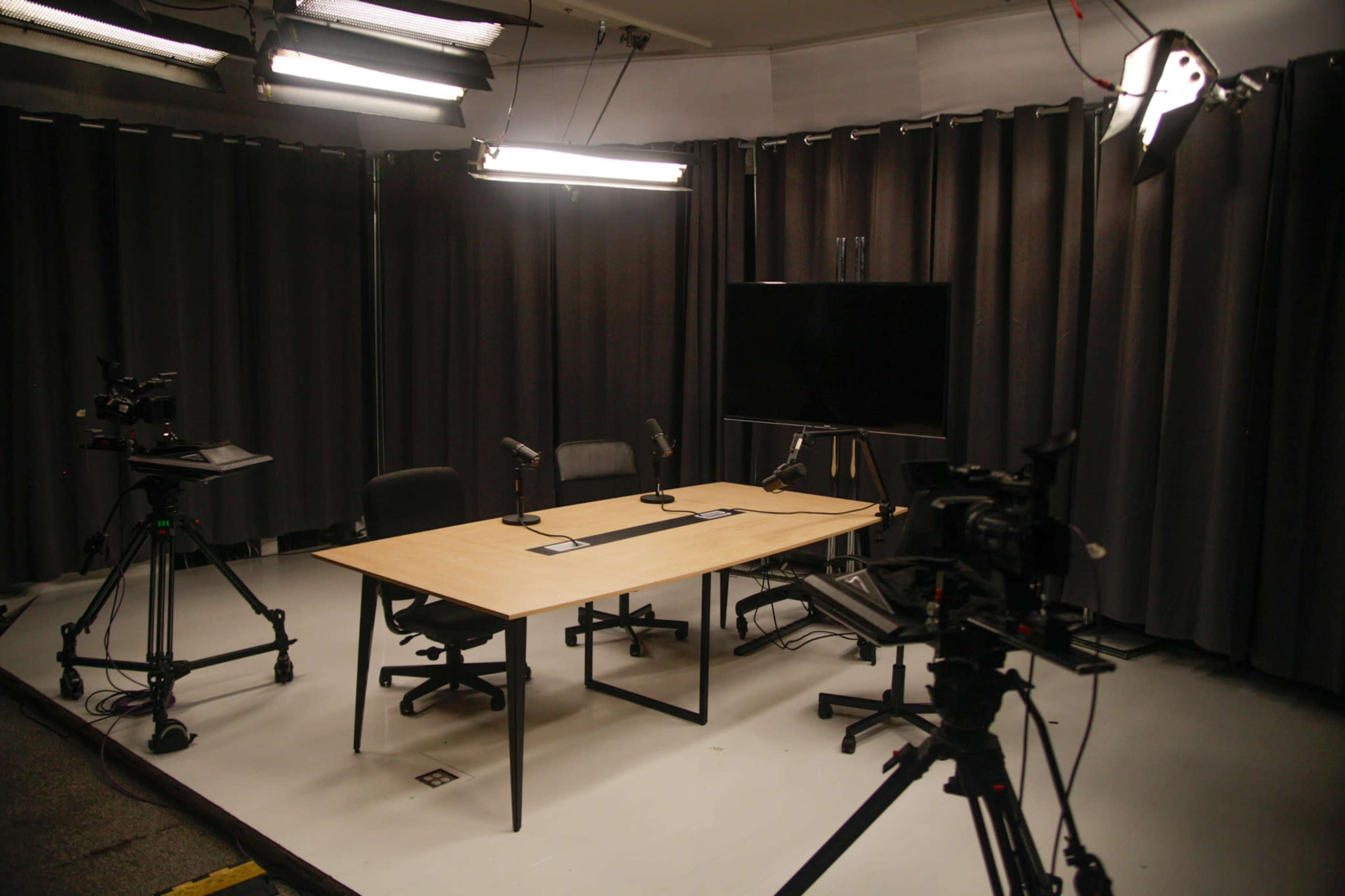 A video recording studio is set up with a table, two chairs, microphones, and various cameras aimed at the scene.