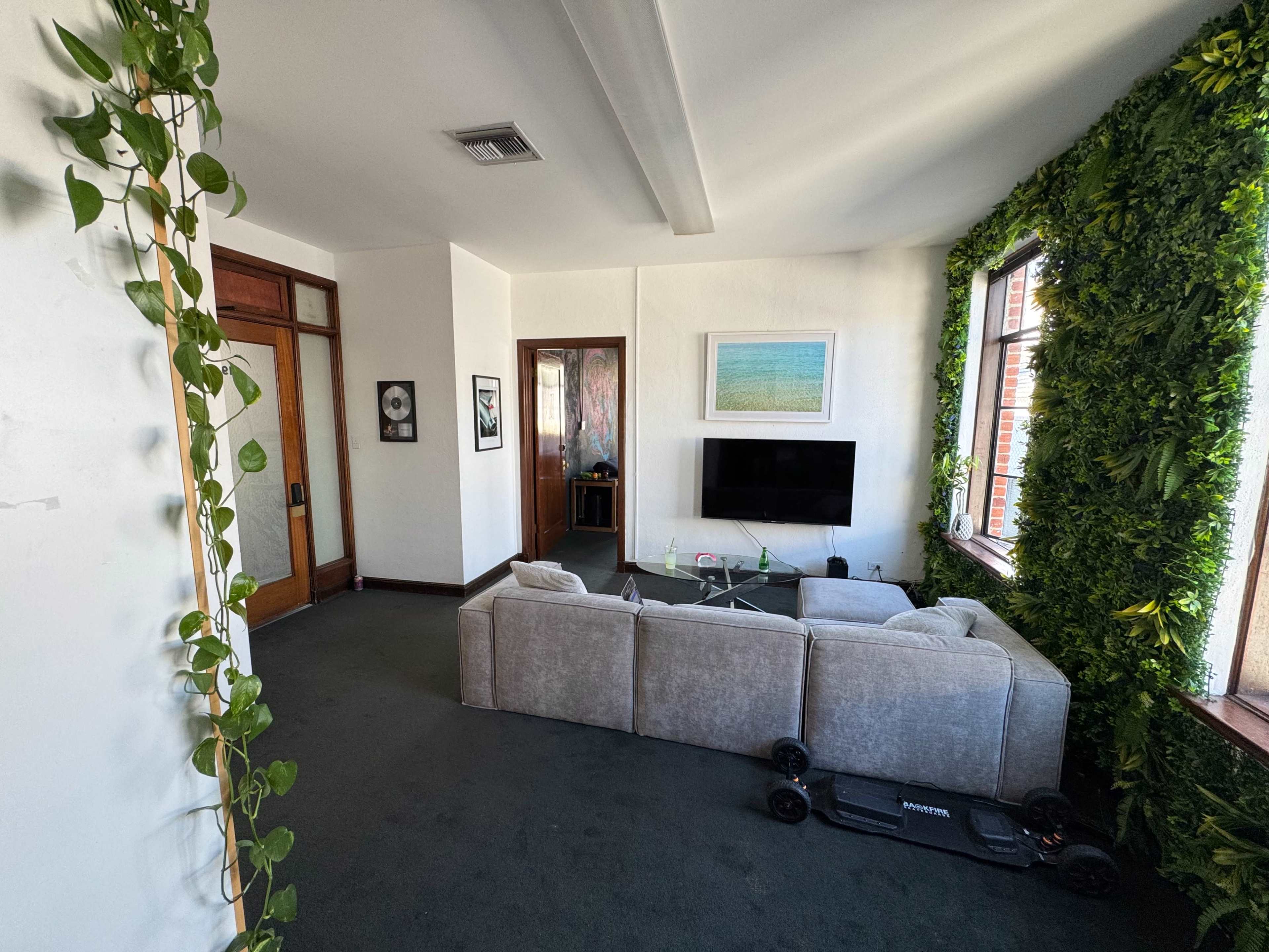 The image shows a modern living room with a grey sectional sofa, a coffee table, and a television, surrounded by decorative plants on the walls.