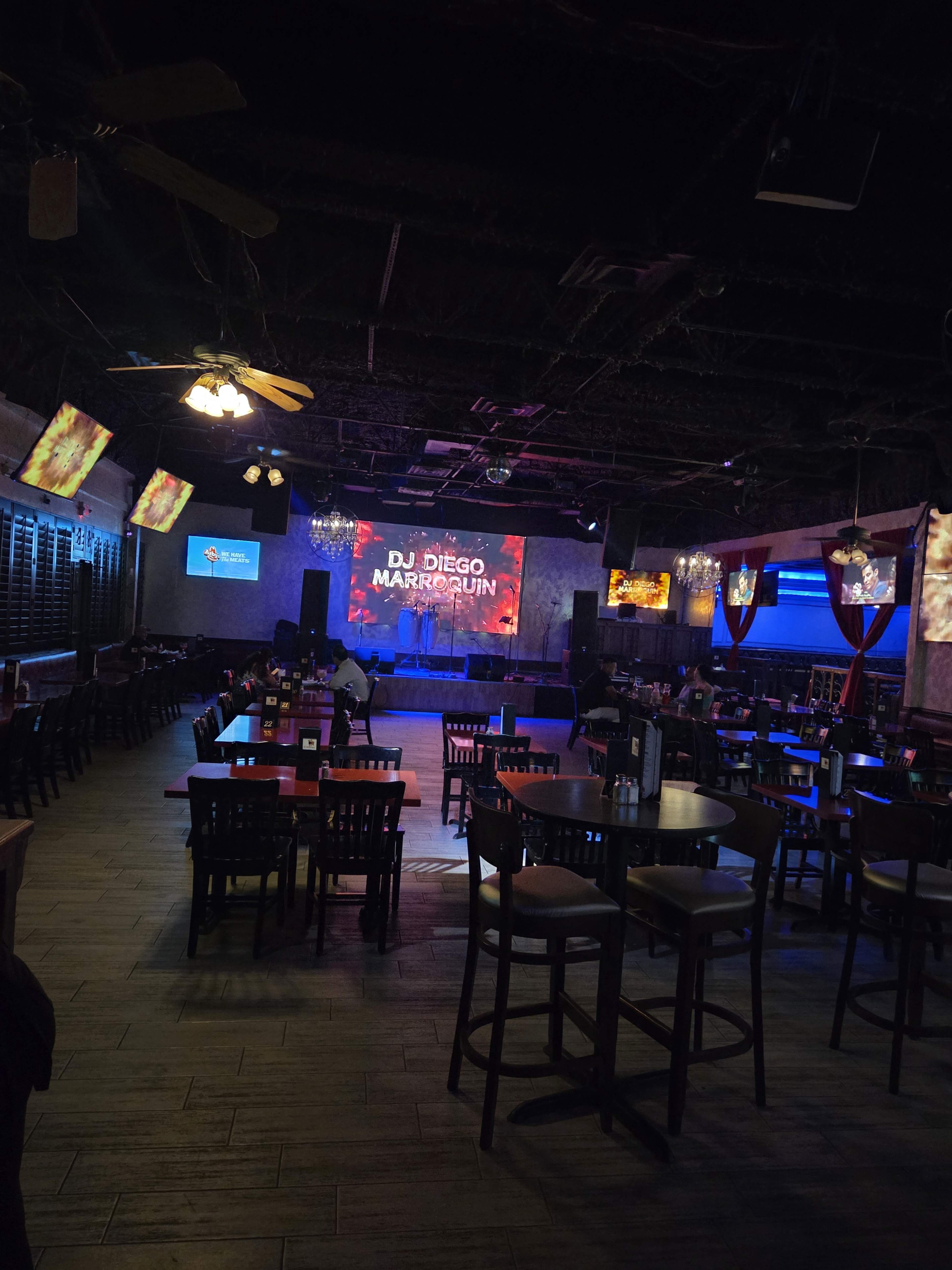 The image shows an empty venue with tables and chairs arranged, a stage with a lighted backdrop, and screens displaying information about an event featuring DJ Diego.