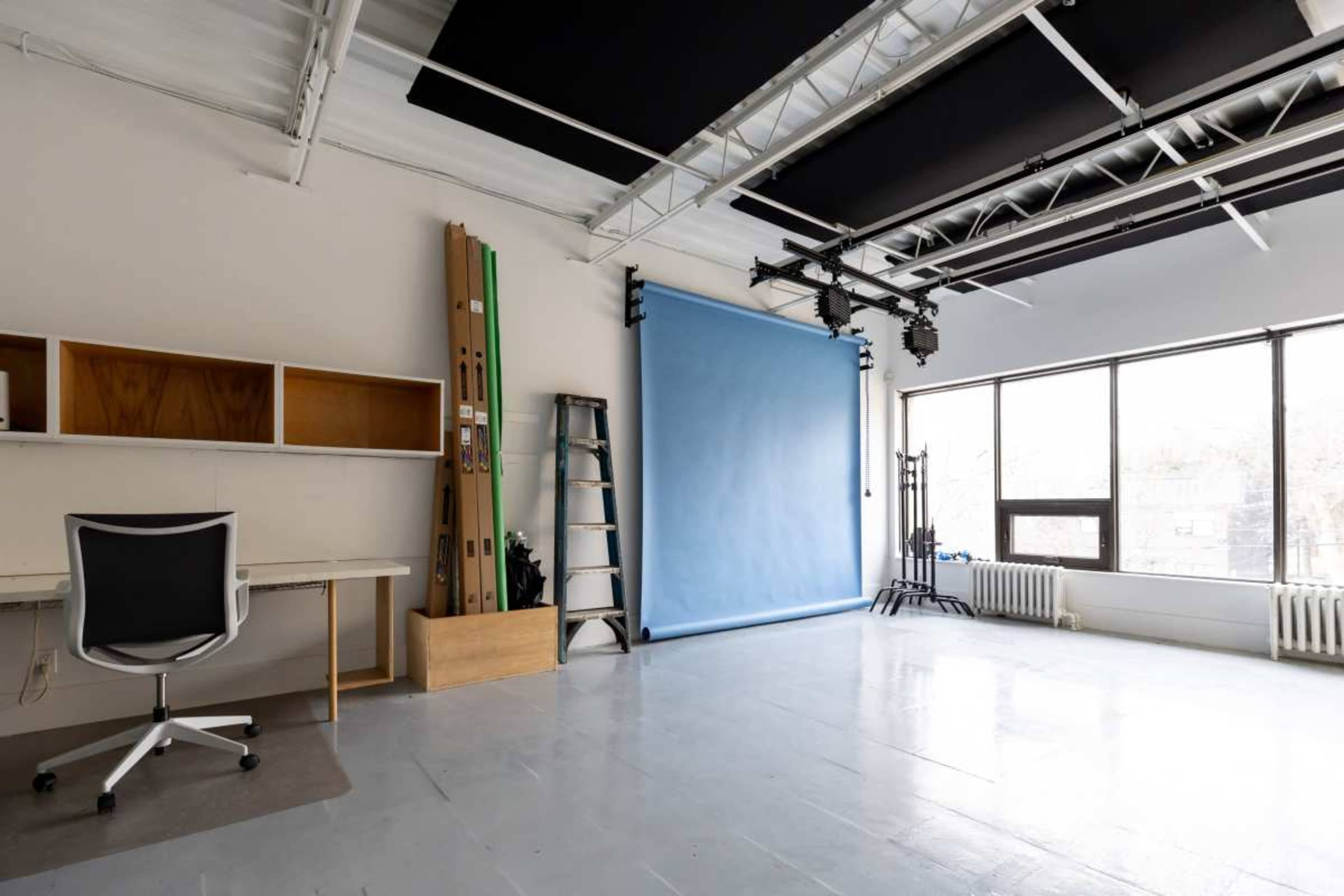 The image shows a minimalist studio space featuring a light blue backdrop, a desk with a chair, a ladder, and large windows allowing natural light to enter.