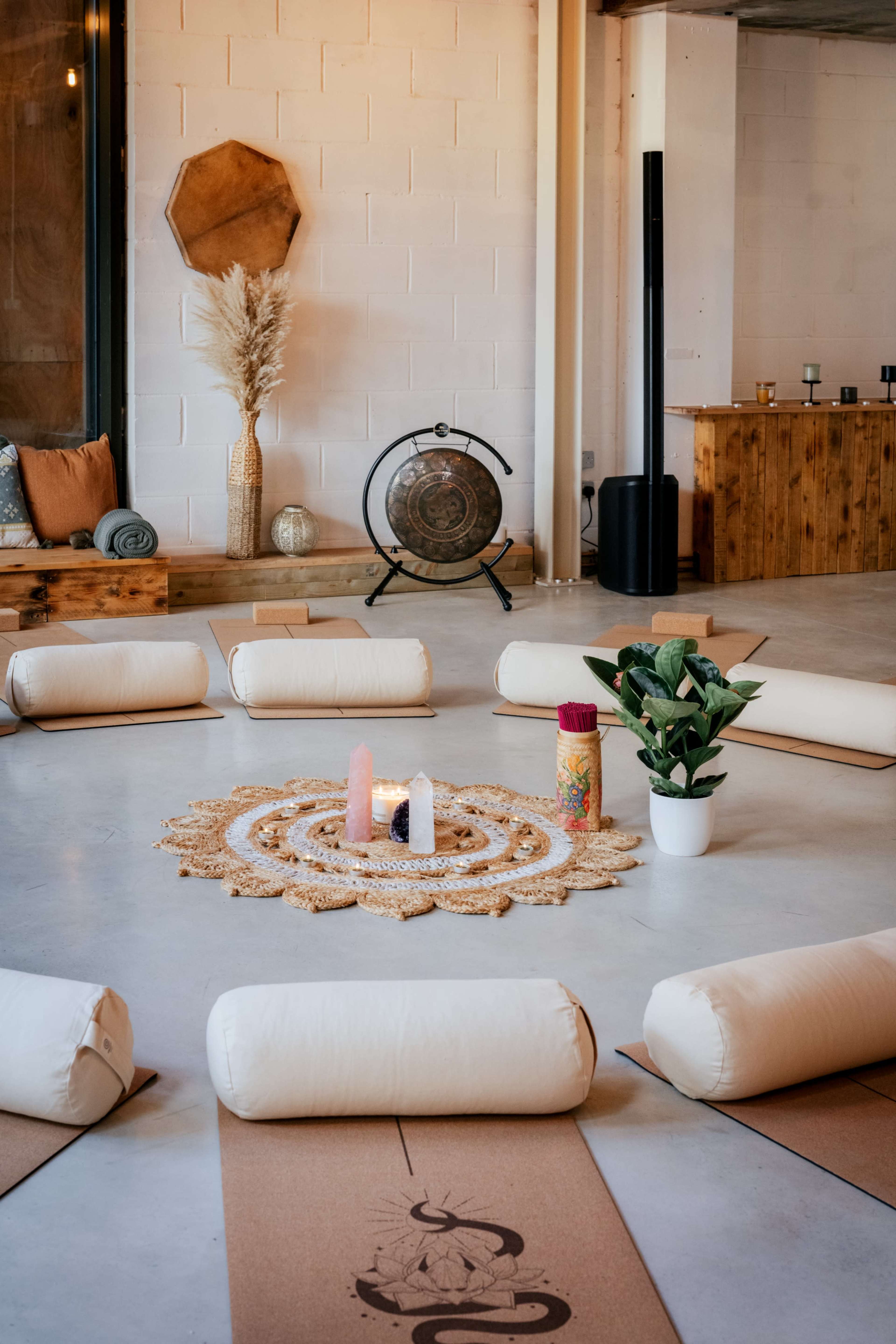 The image shows a meditation room arranged in a circular setup with cushions, a decorative centerpiece made of natural materials, candles, and a plant in a pot.