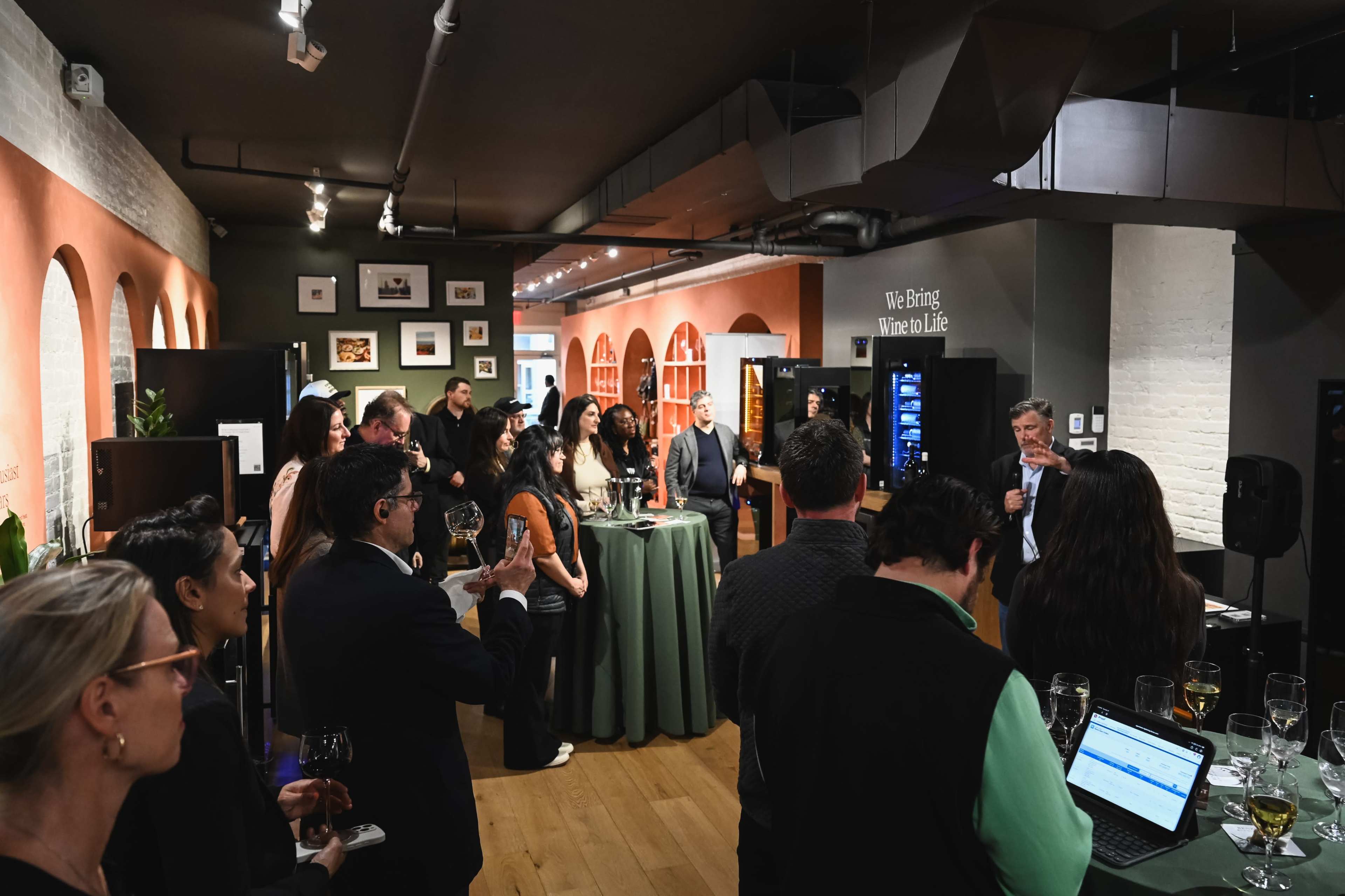 A group of people mingles and listens attentively in a wine-tasting event held in a stylish venue with arches and soft lighting.