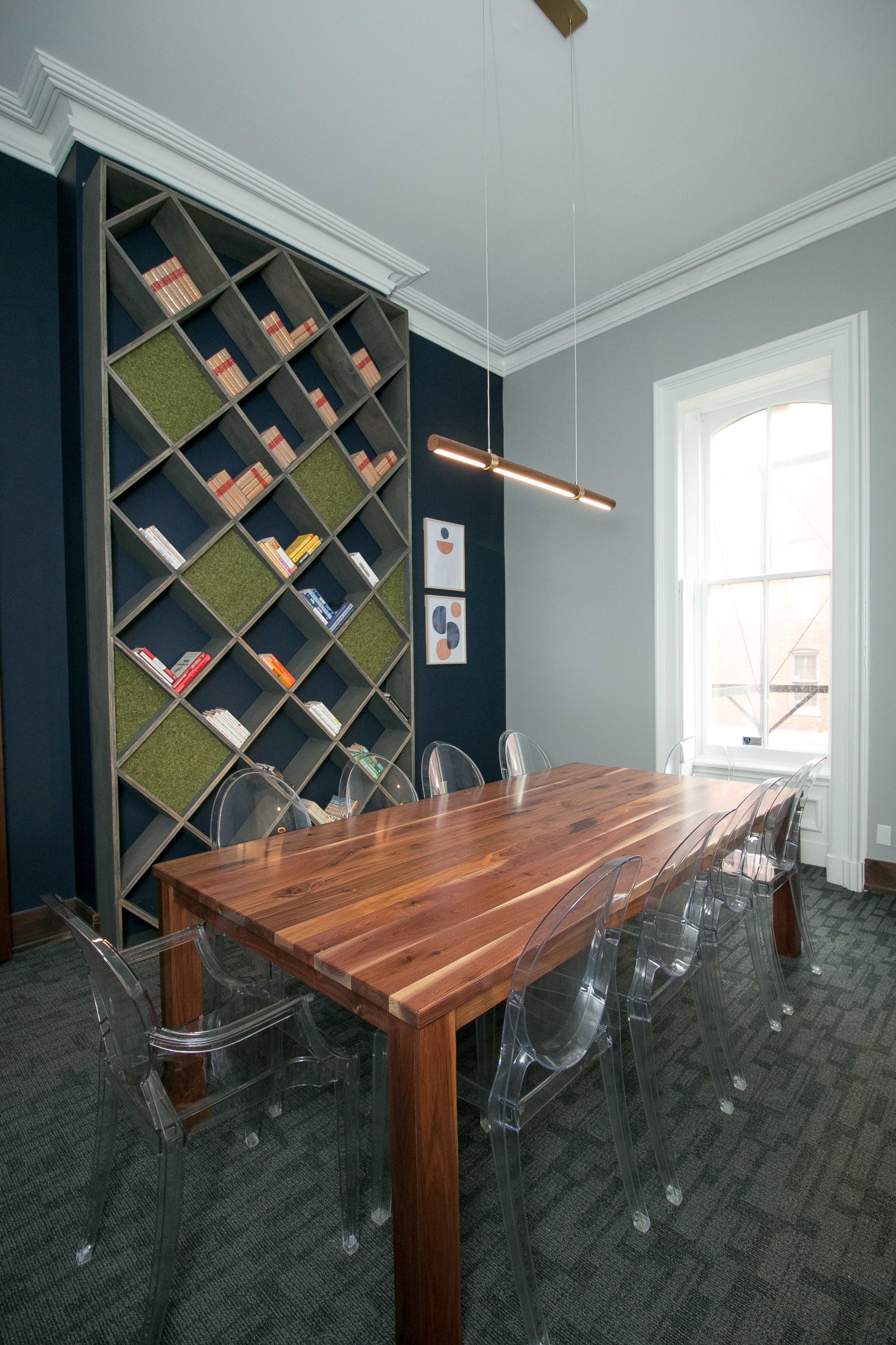 A modern meeting room featuring a large wooden table surrounded by transparent chairs, with a geometric bookshelf filled with books and decorative elements in the background.