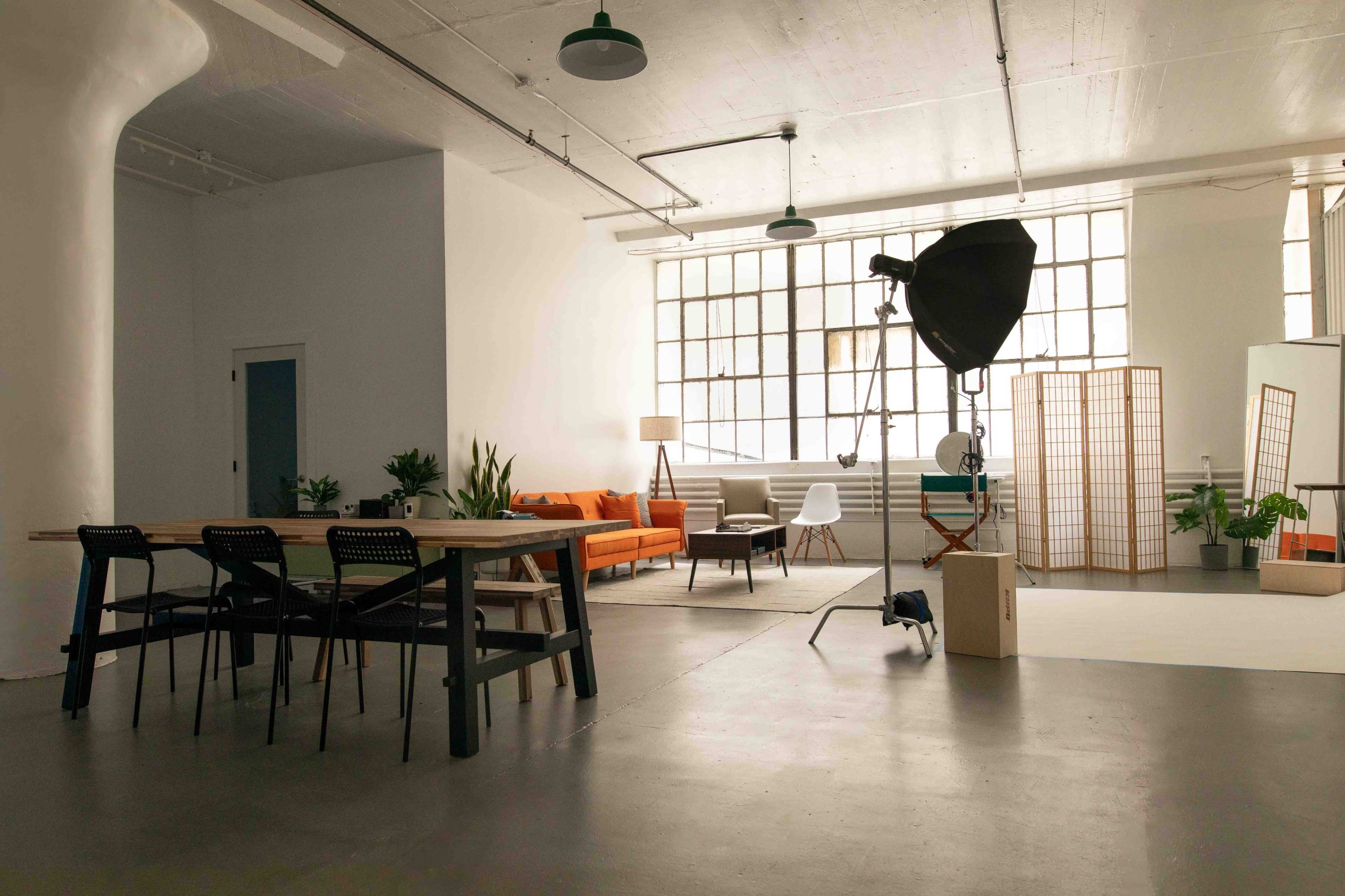A spacious, modern studio with a dining area, a living space featuring an orange sofa, and a photography setup illuminated by natural light from large windows.