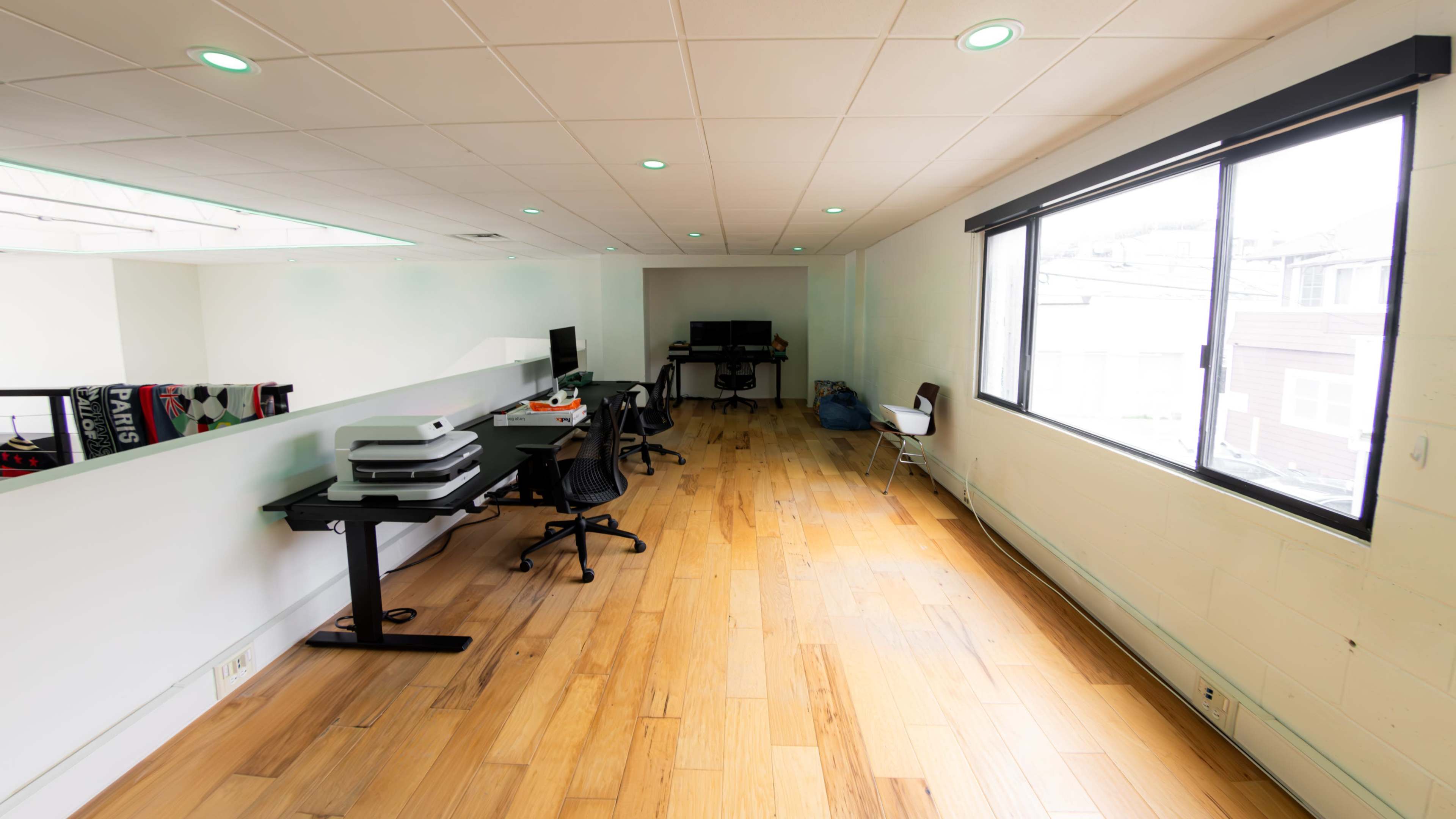 A spacious office area with wooden flooring, equipped with multiple desks, computers, and a large window allowing natural light.