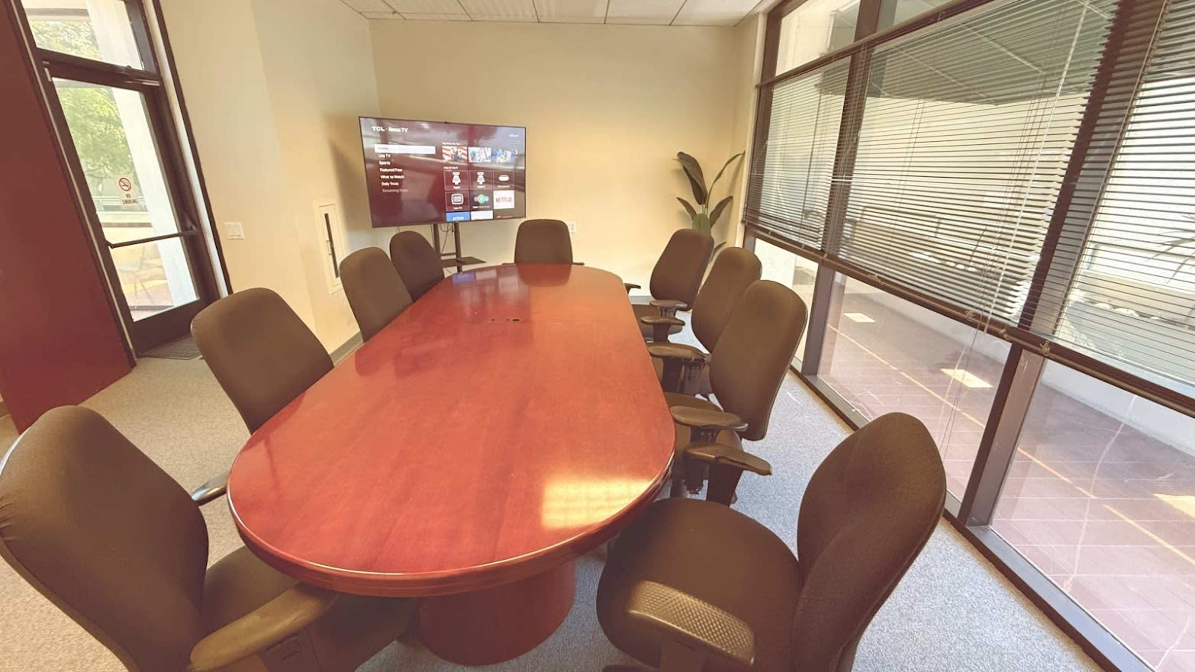The image shows a conference room with a large oval wooden table surrounded by eight black chairs, featuring a television screen mounted on the wall.